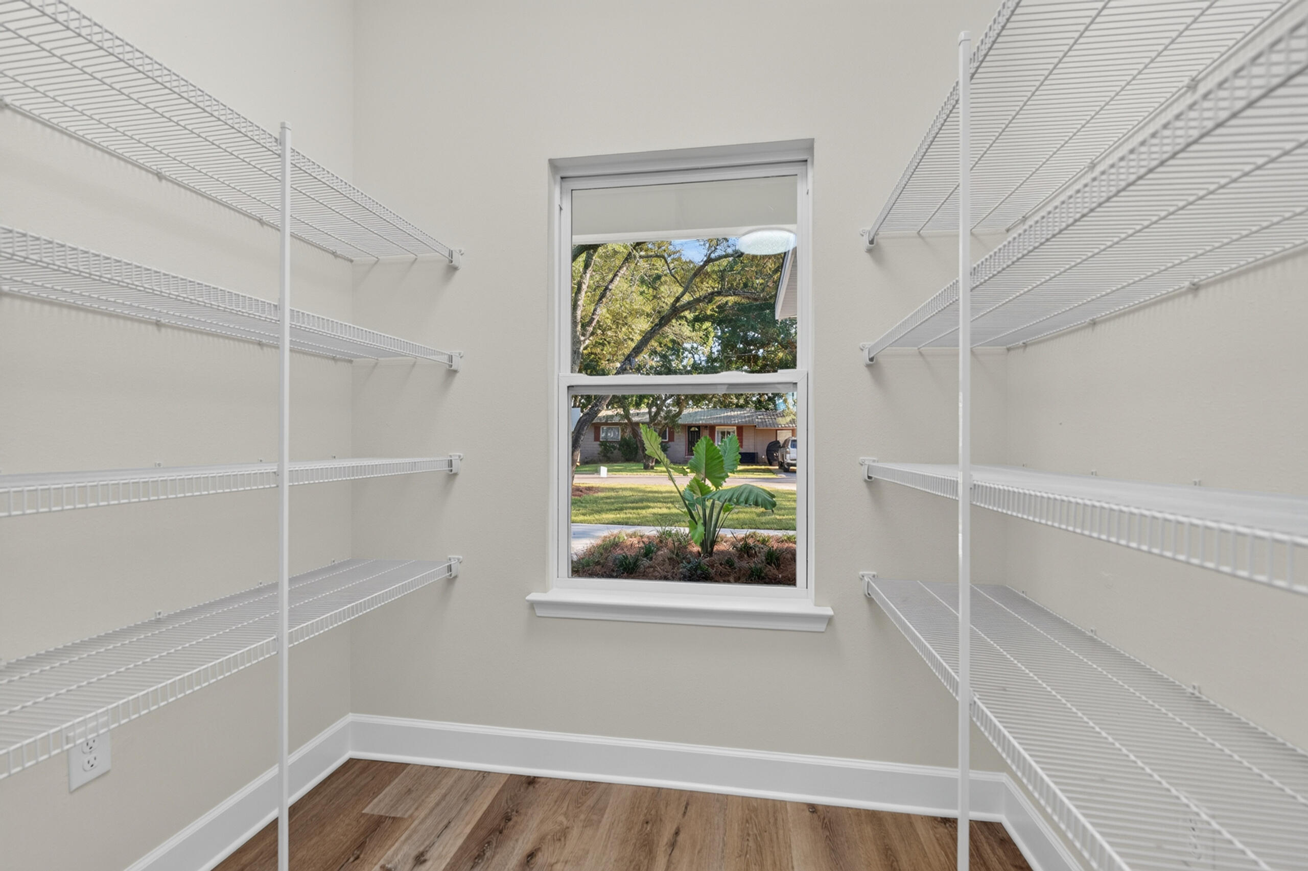 Walk-in closet with white wire shelving, wood-look flooring, and a window overlooking a leafy yard.