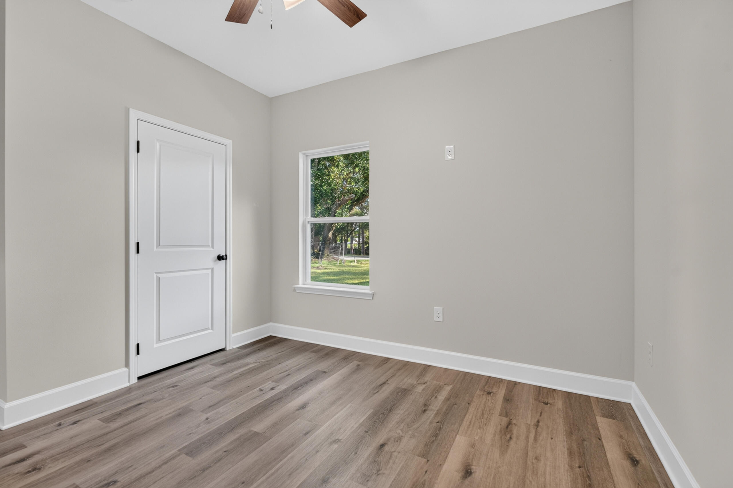 Empty room with wood-look flooring, a white paneled door, and a window overlooking a leafy yard.