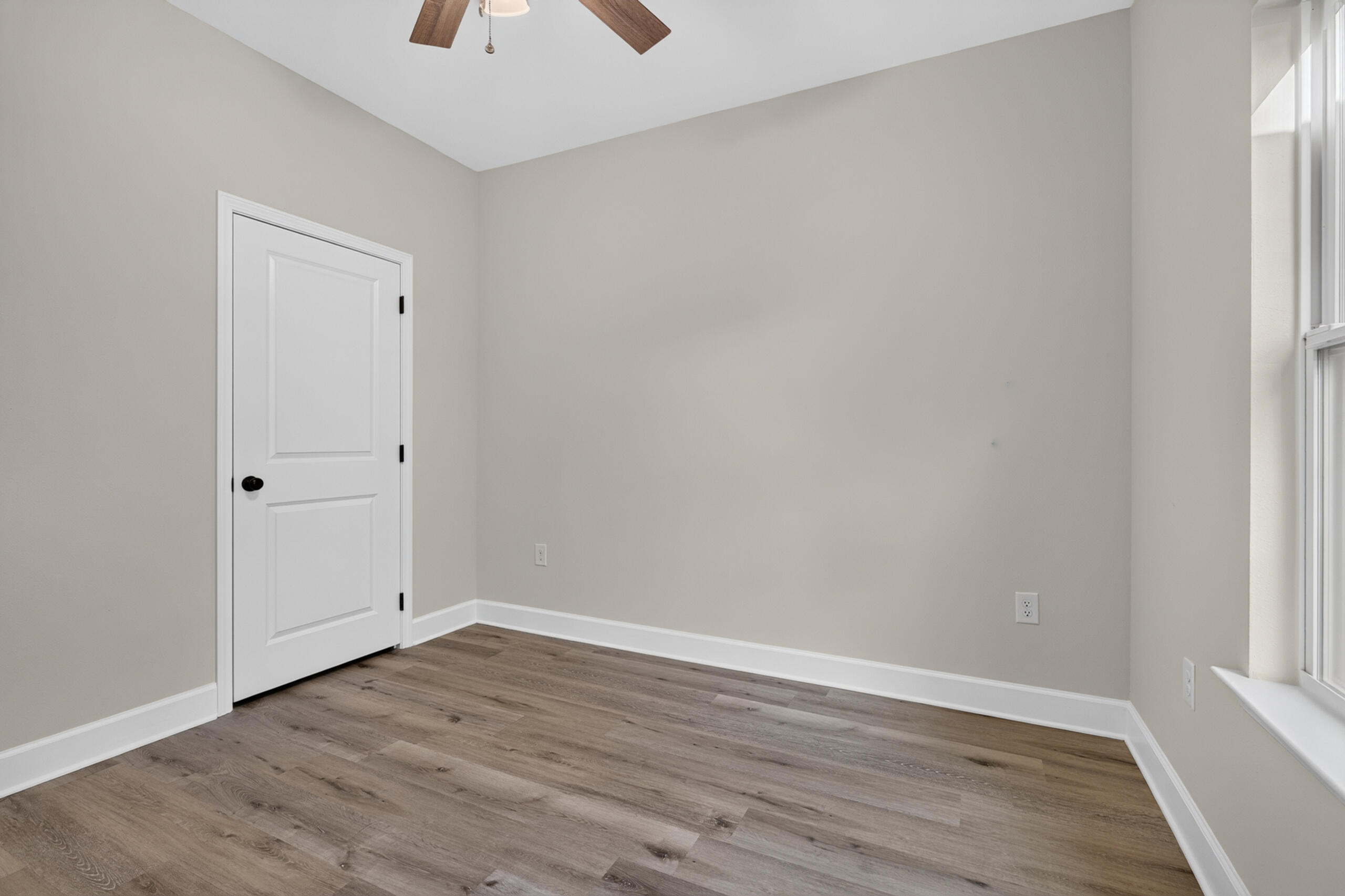 Vacant room with wood-look flooring, a paneled door, ceiling fan, and large window.