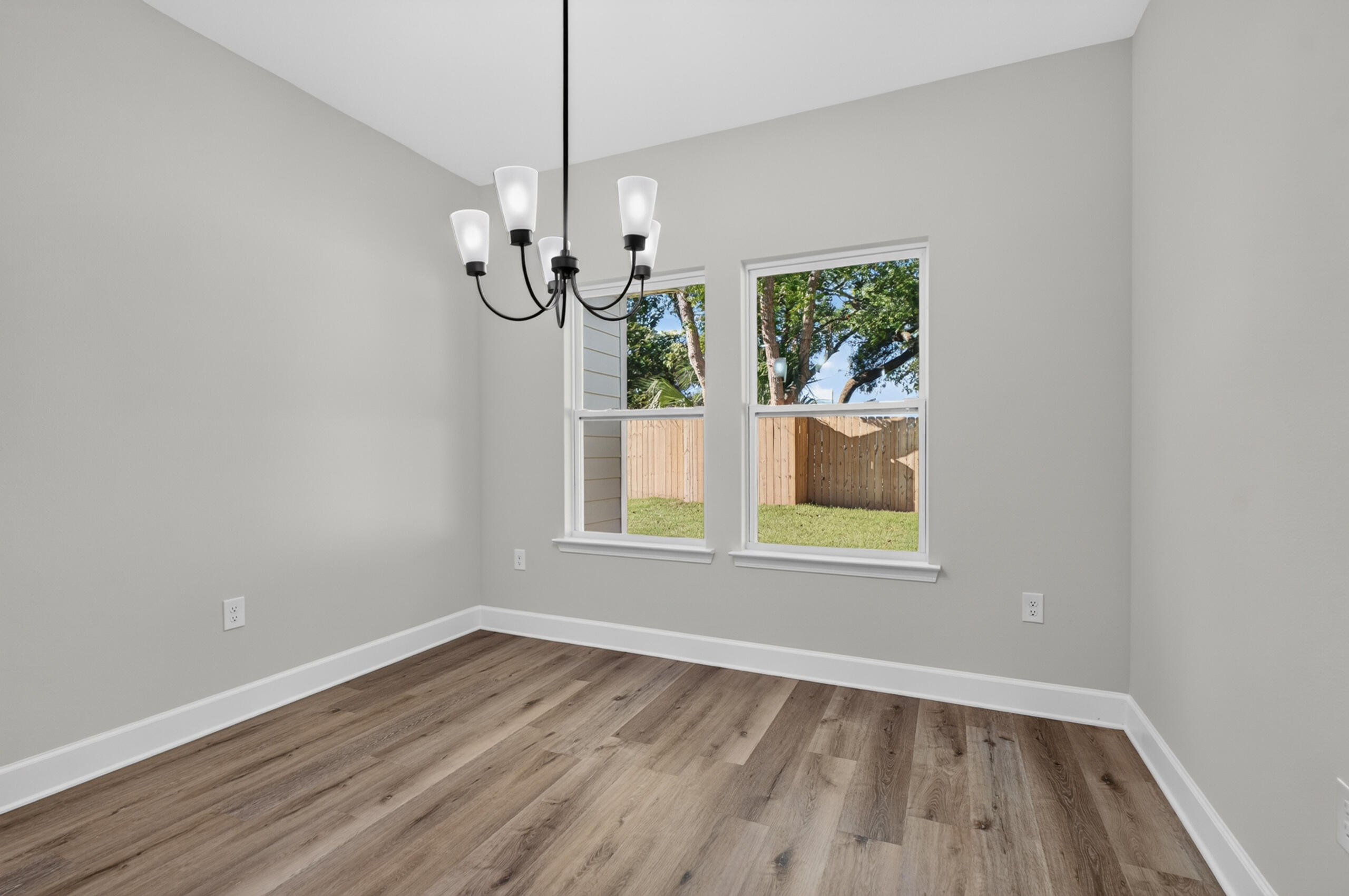 Empty room with wood-look flooring, a black chandelier, and two windows facing a fenced yard.