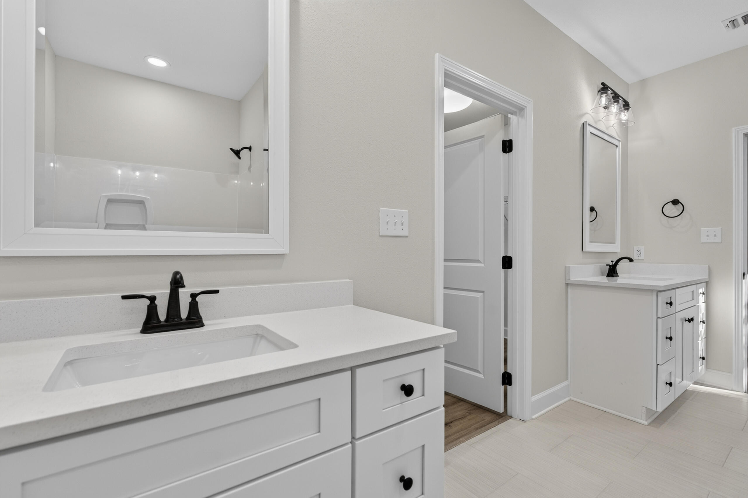 Bathroom with two white vanities, black fixtures, large mirrors, and light tile flooring.