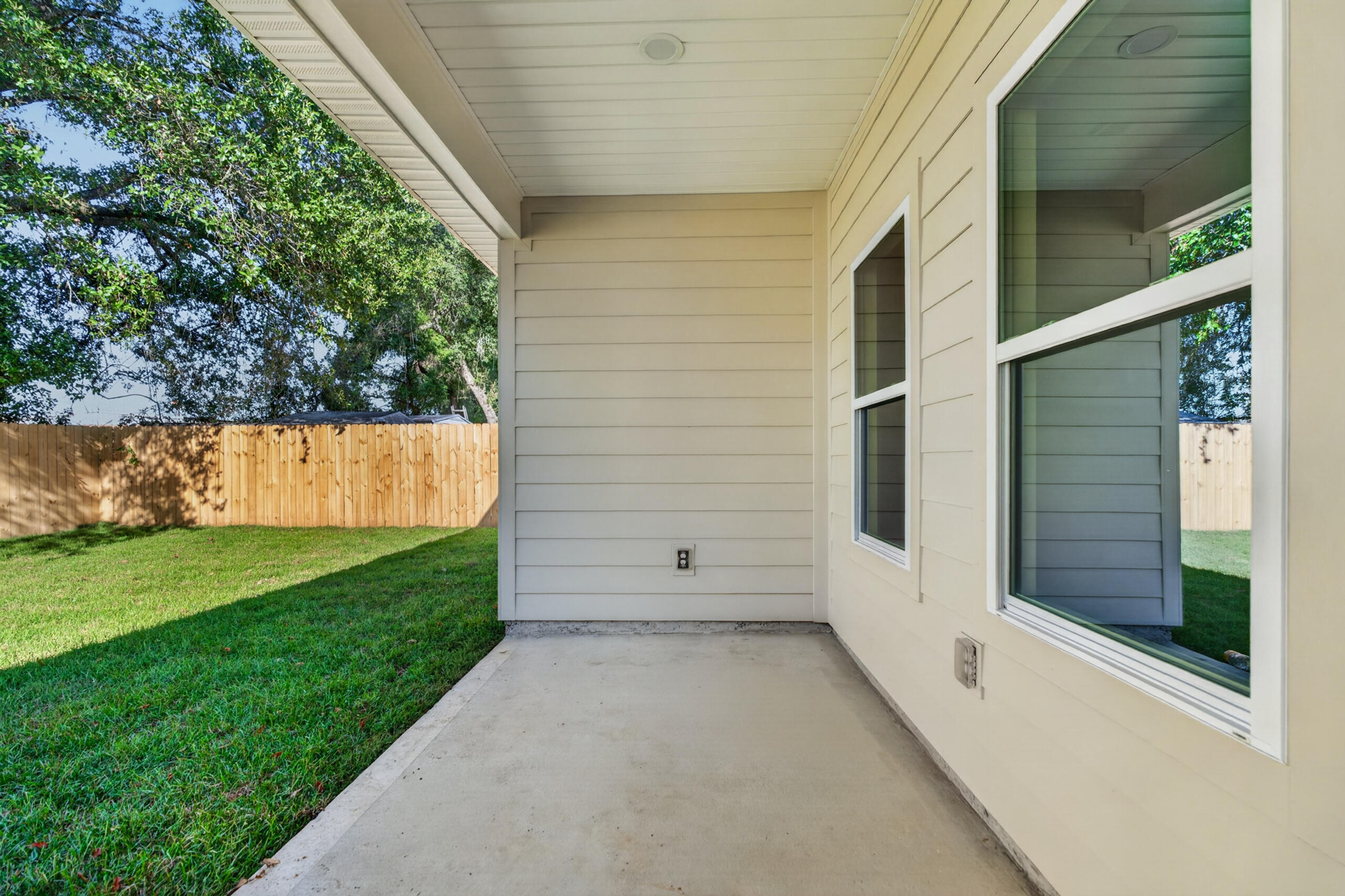 Covered concrete patio beside the home, overlooking a fenced grassy yard with mature trees.