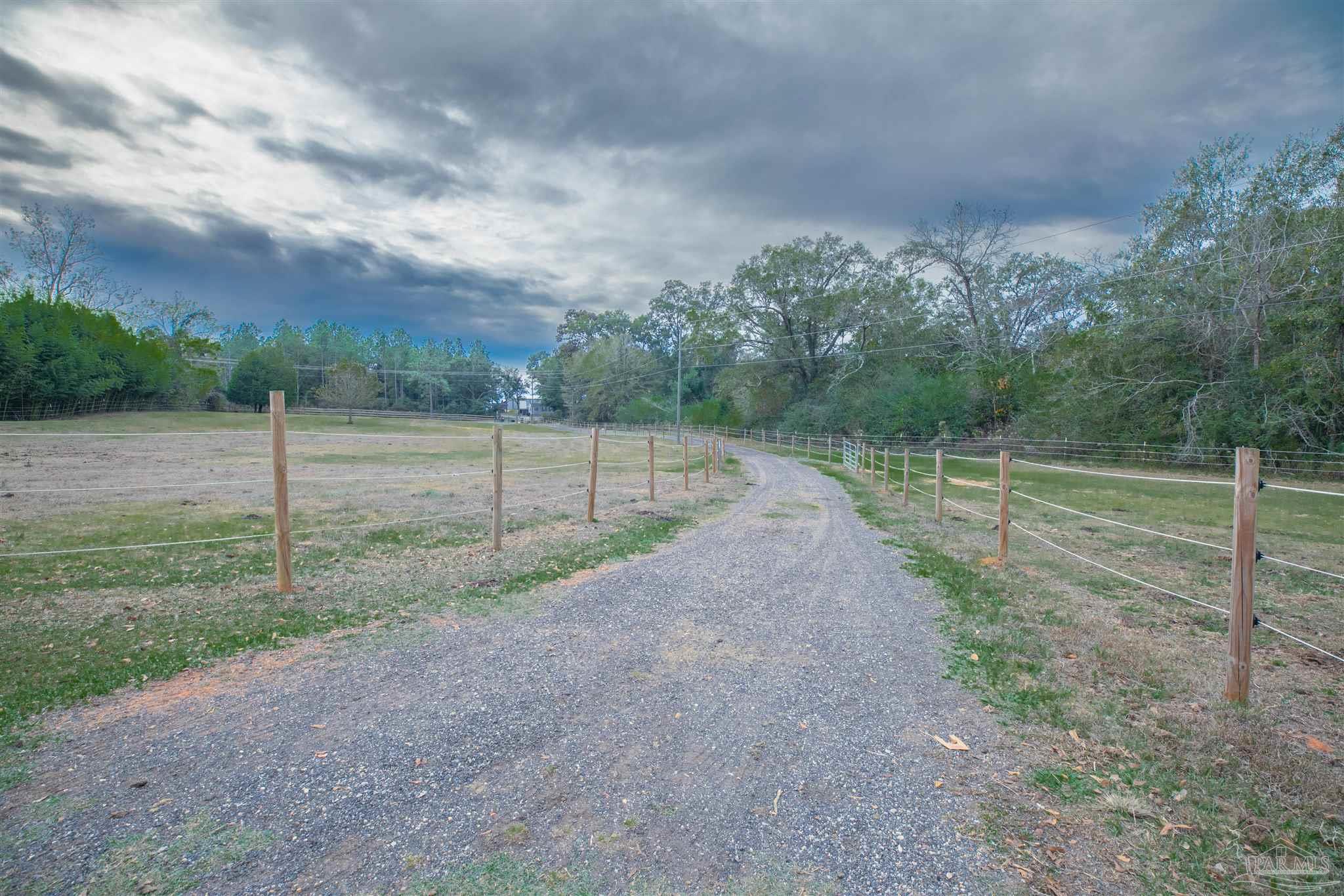 Gravel driveway curves between fenced grassy paddocks toward a wooded property.