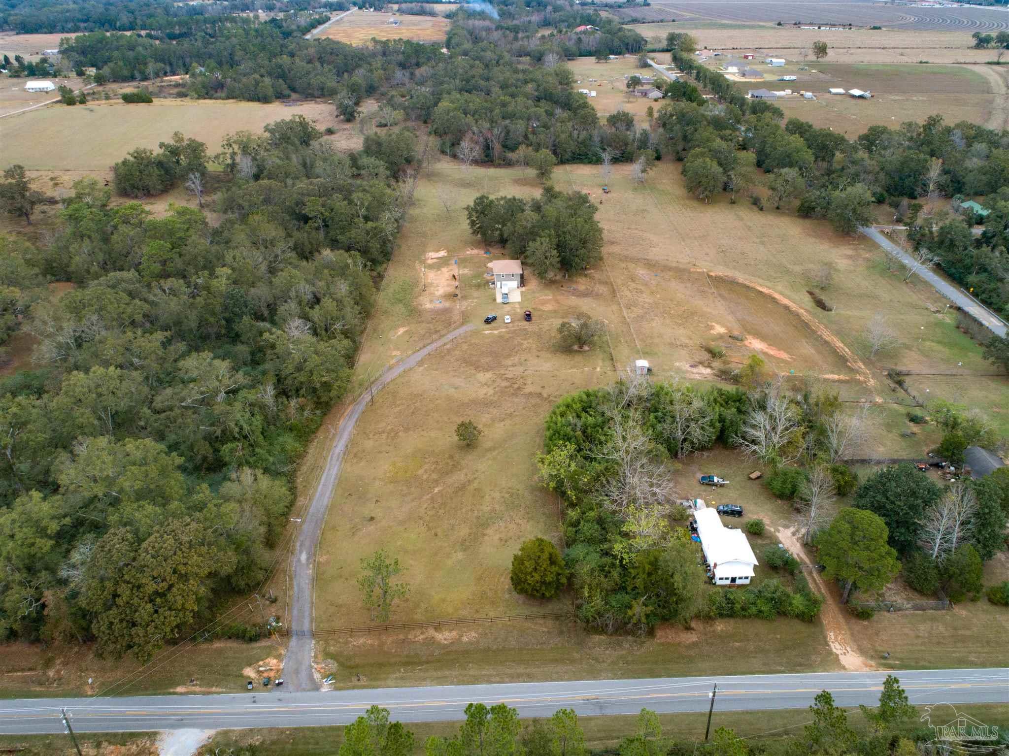 Aerial view of rural acreage with two homes, a curved driveway, open fields, and surrounding woods.