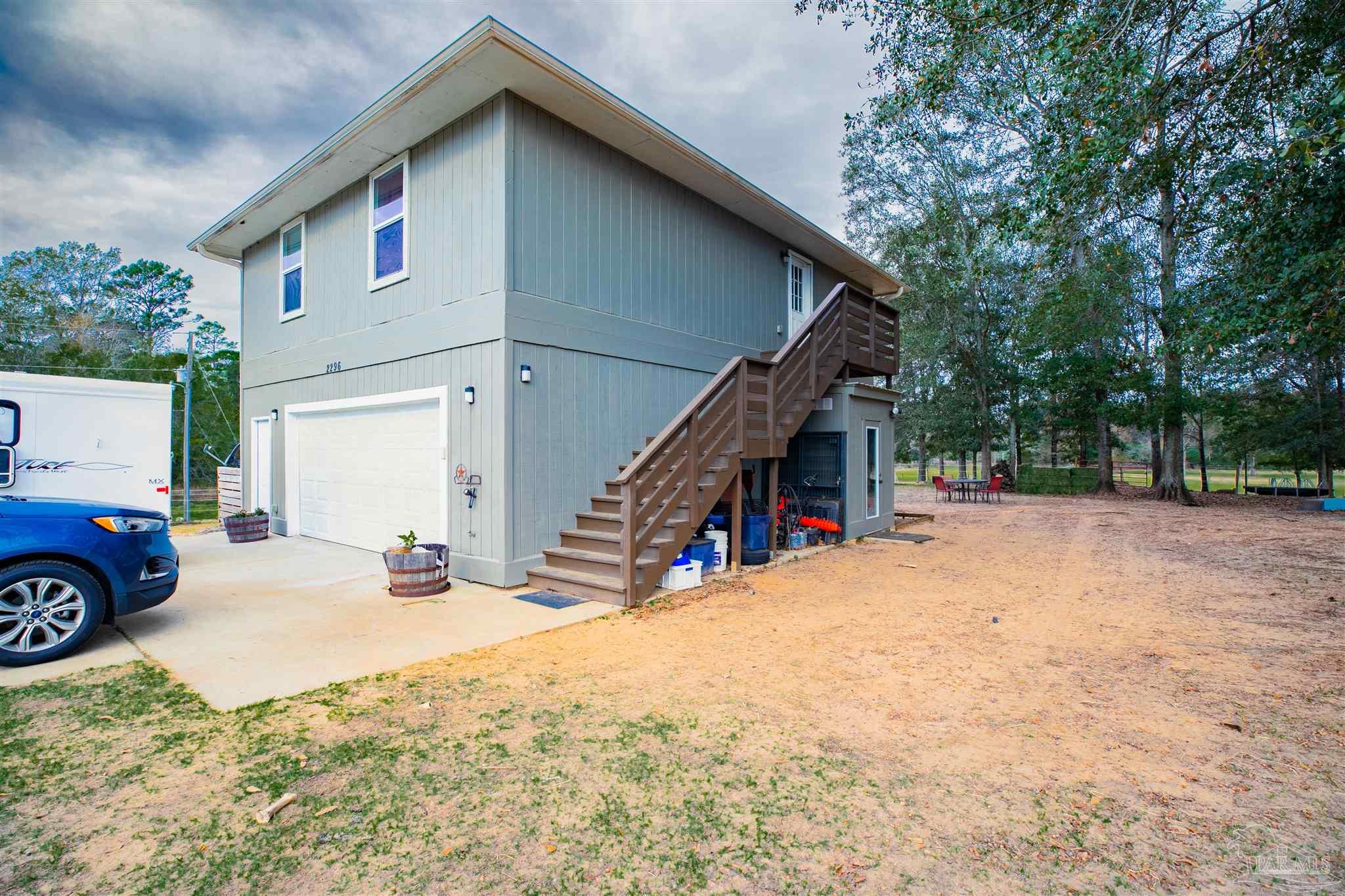 Gray two-story home with a garage, exterior staircase, sandy yard, and mature trees.