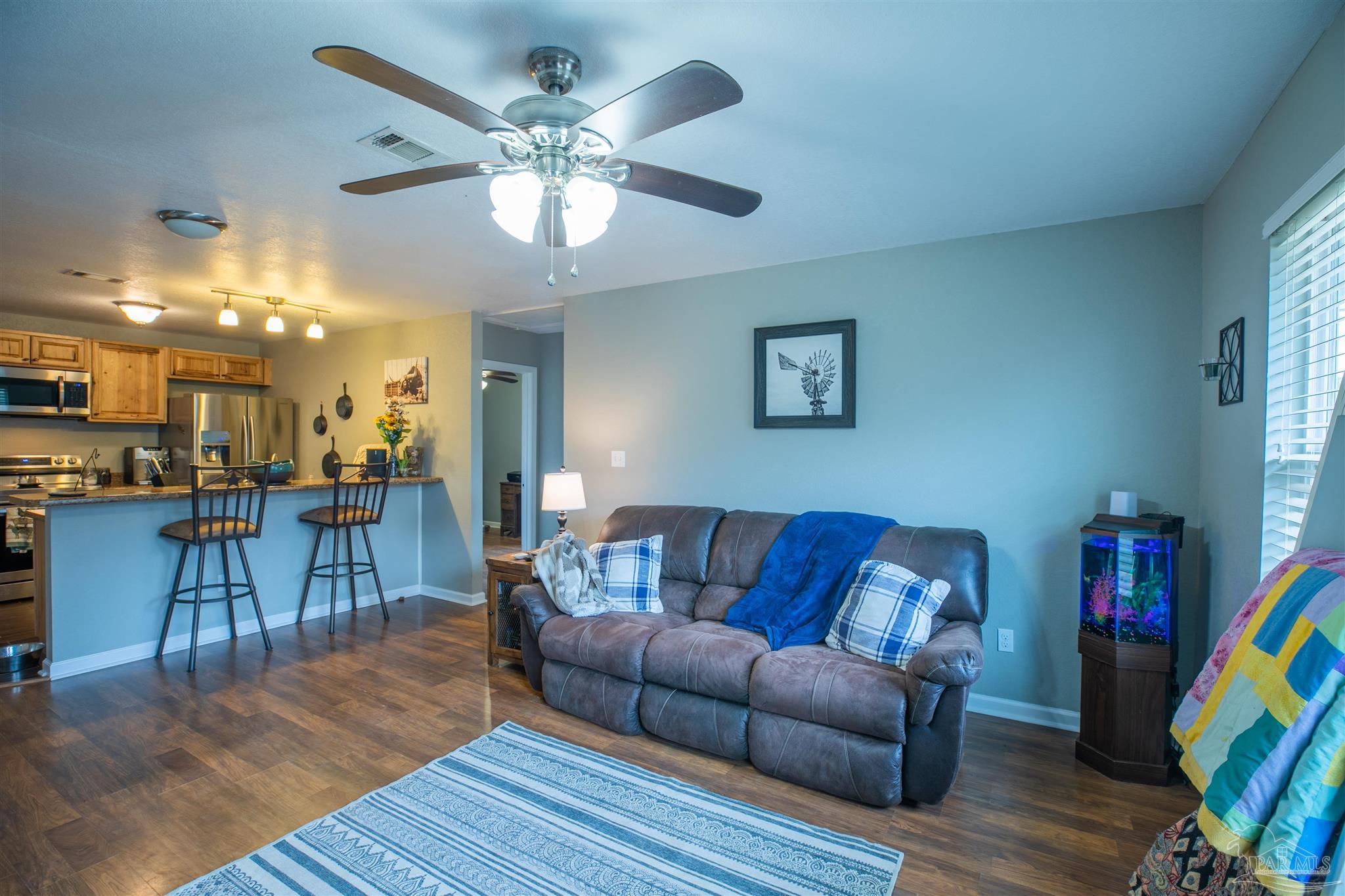 Living room with gray reclining sofa and ceiling fan, open to a kitchen with a breakfast bar.