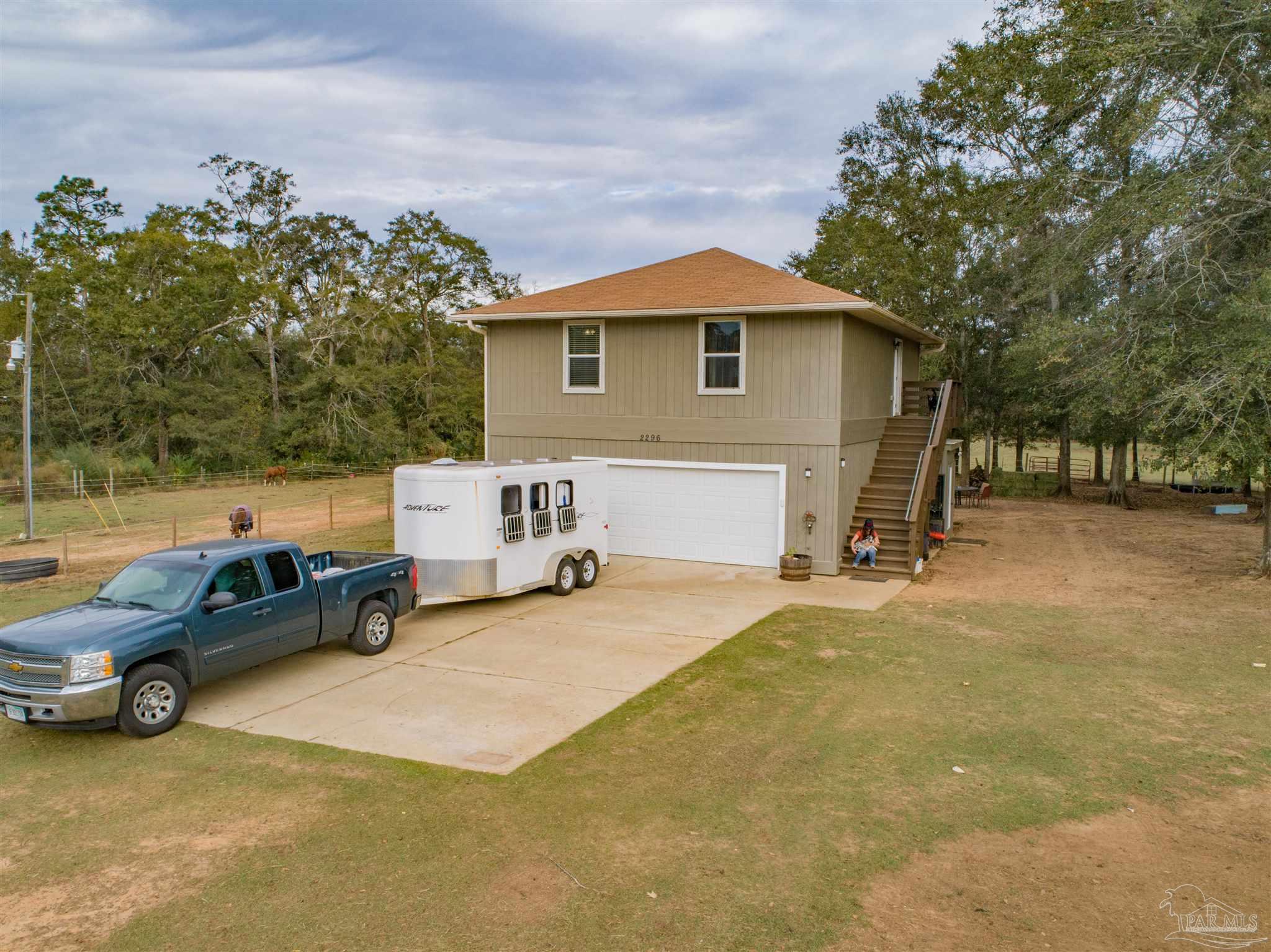 Raised beige home with exterior staircase, garage, driveway, pickup truck, and horse trailer.