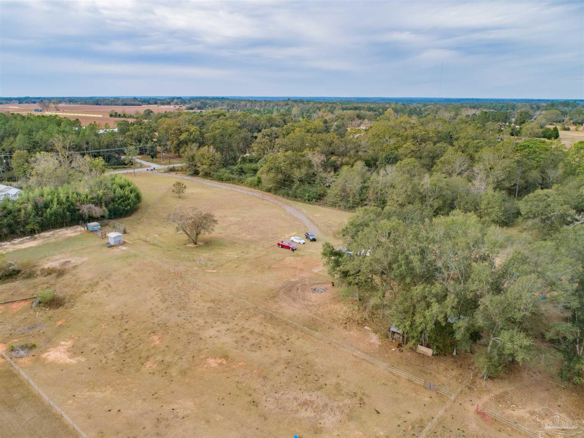 Aerial view of fenced pastureland bordered by woods, with a curving driveway and small outbuildings.
