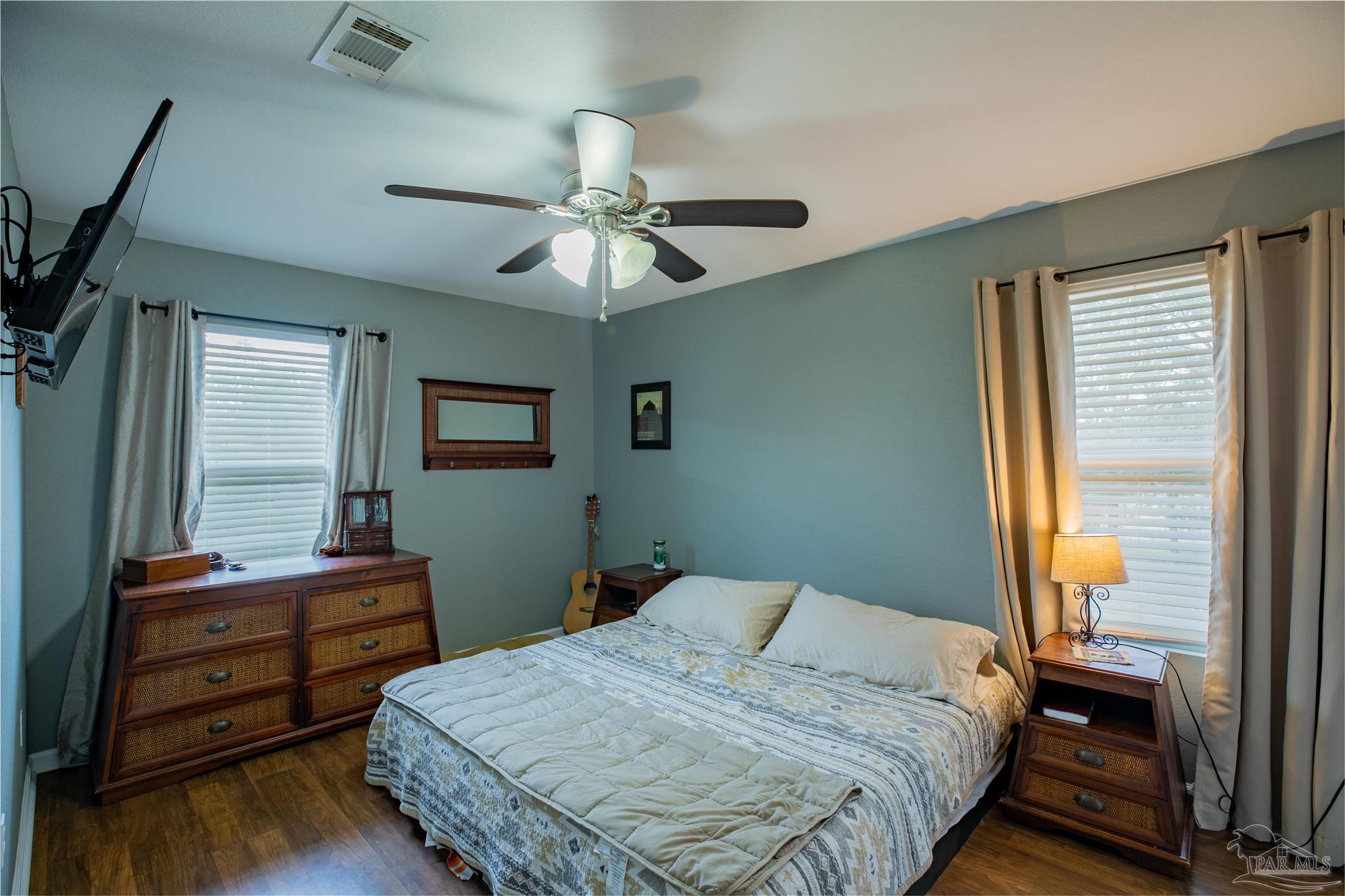 Furnished bedroom with a patterned bedspread, dark wood furniture, two curtained windows, and a ceiling fan.
