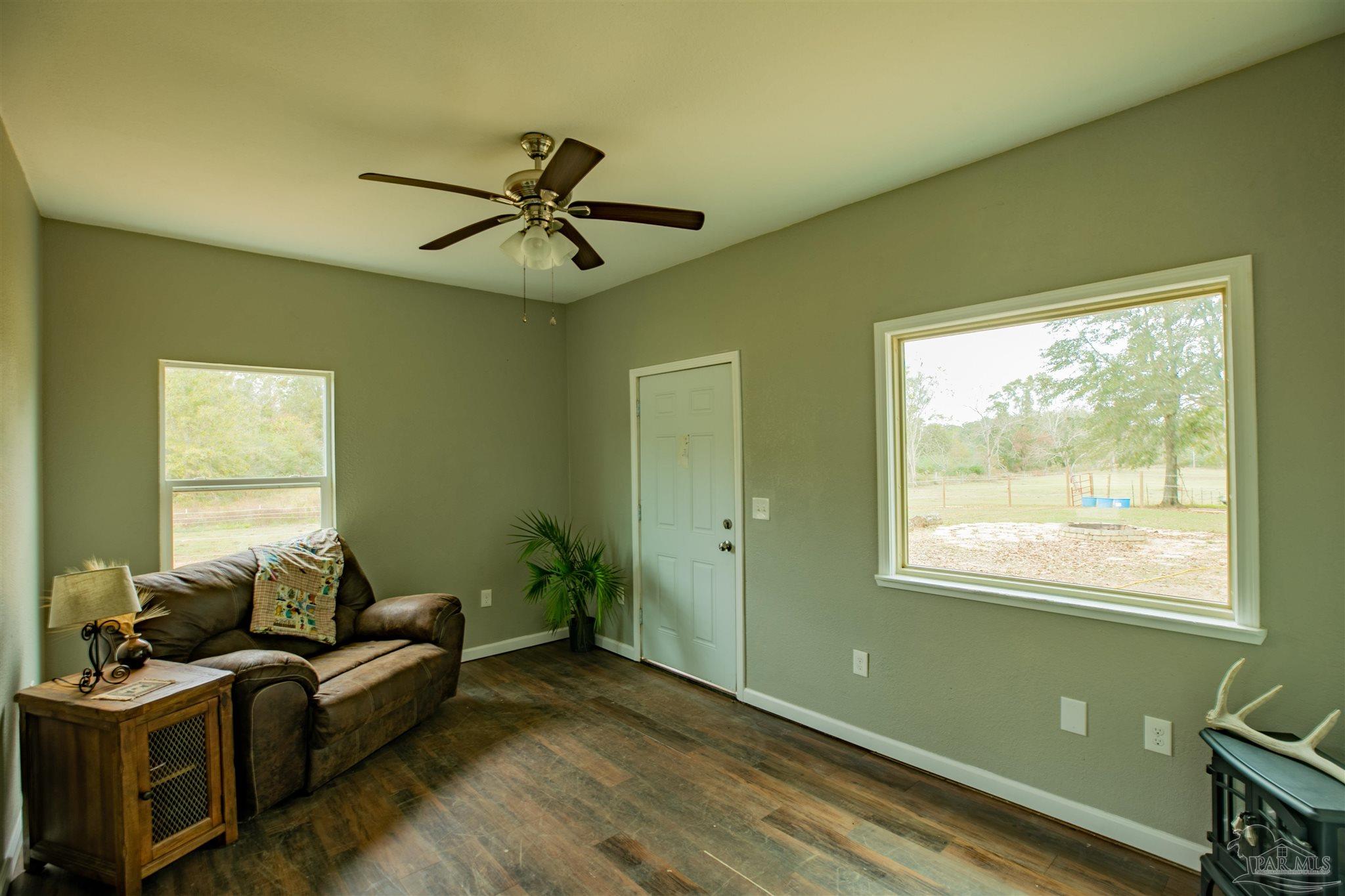 Living room with a brown sofa, ceiling fan, and large window overlooking a fenced yard.