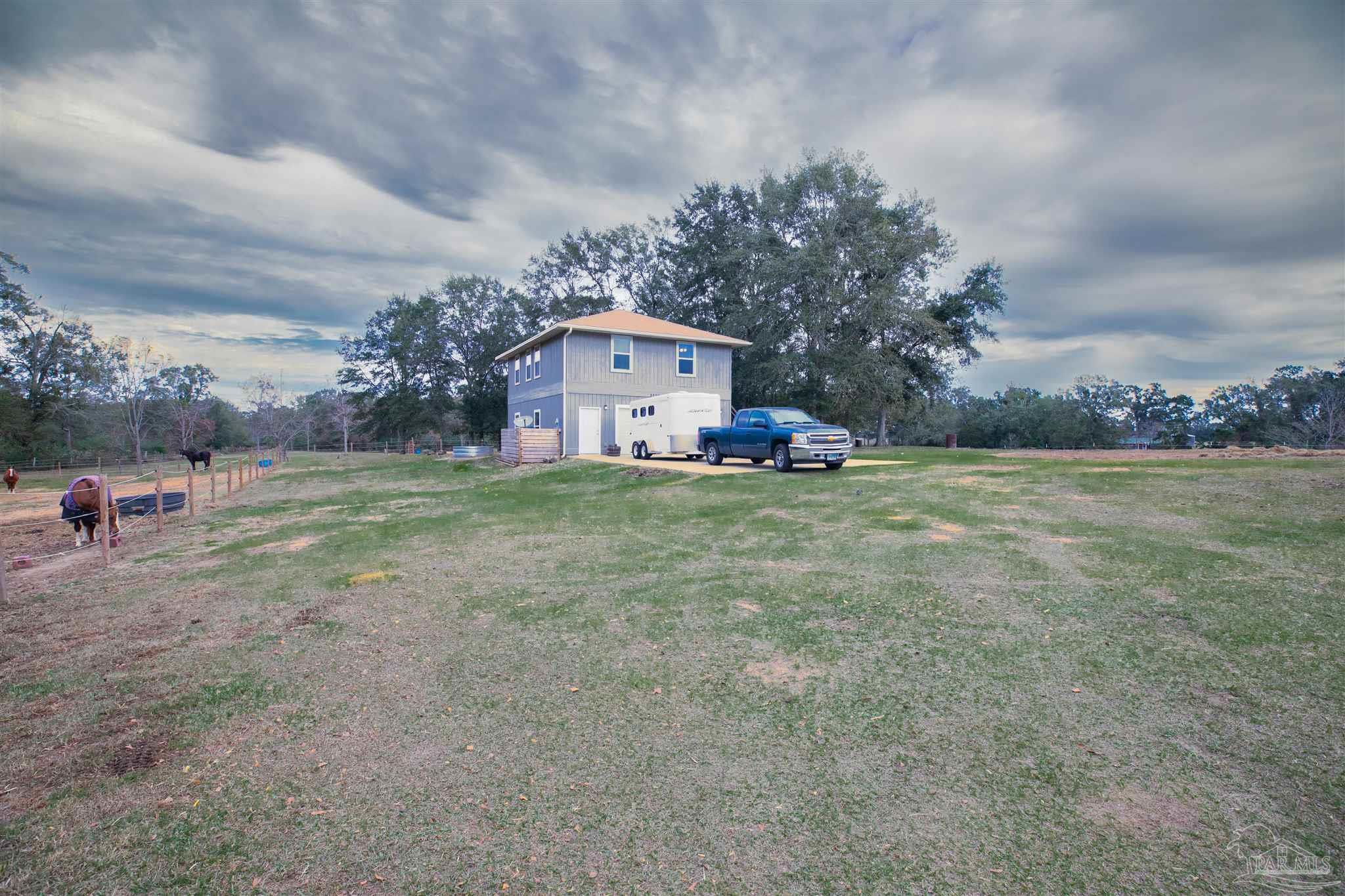 Rural property with a two-story outbuilding, blue pickup and trailer, fenced paddock, and open grassy yard.