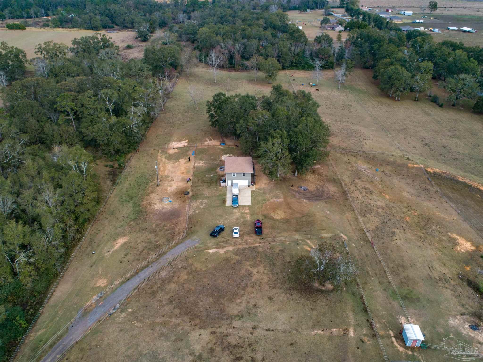 Aerial view of a rural property with a small house, fenced pasture, mature trees, and a curved driveway.