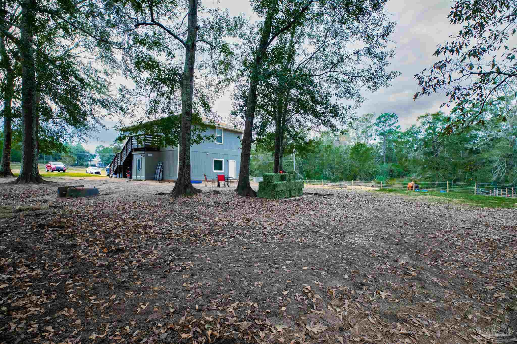 Gray two-story home beneath mature trees, with an exterior staircase and leaf-covered yard.