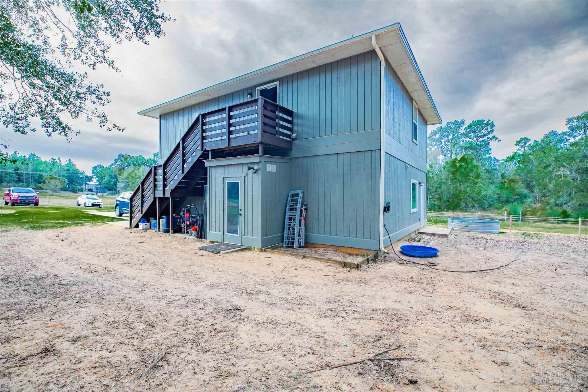 Gray two-story home with a raised balcony and exterior stairs beside a sandy yard and stock-tank pool.