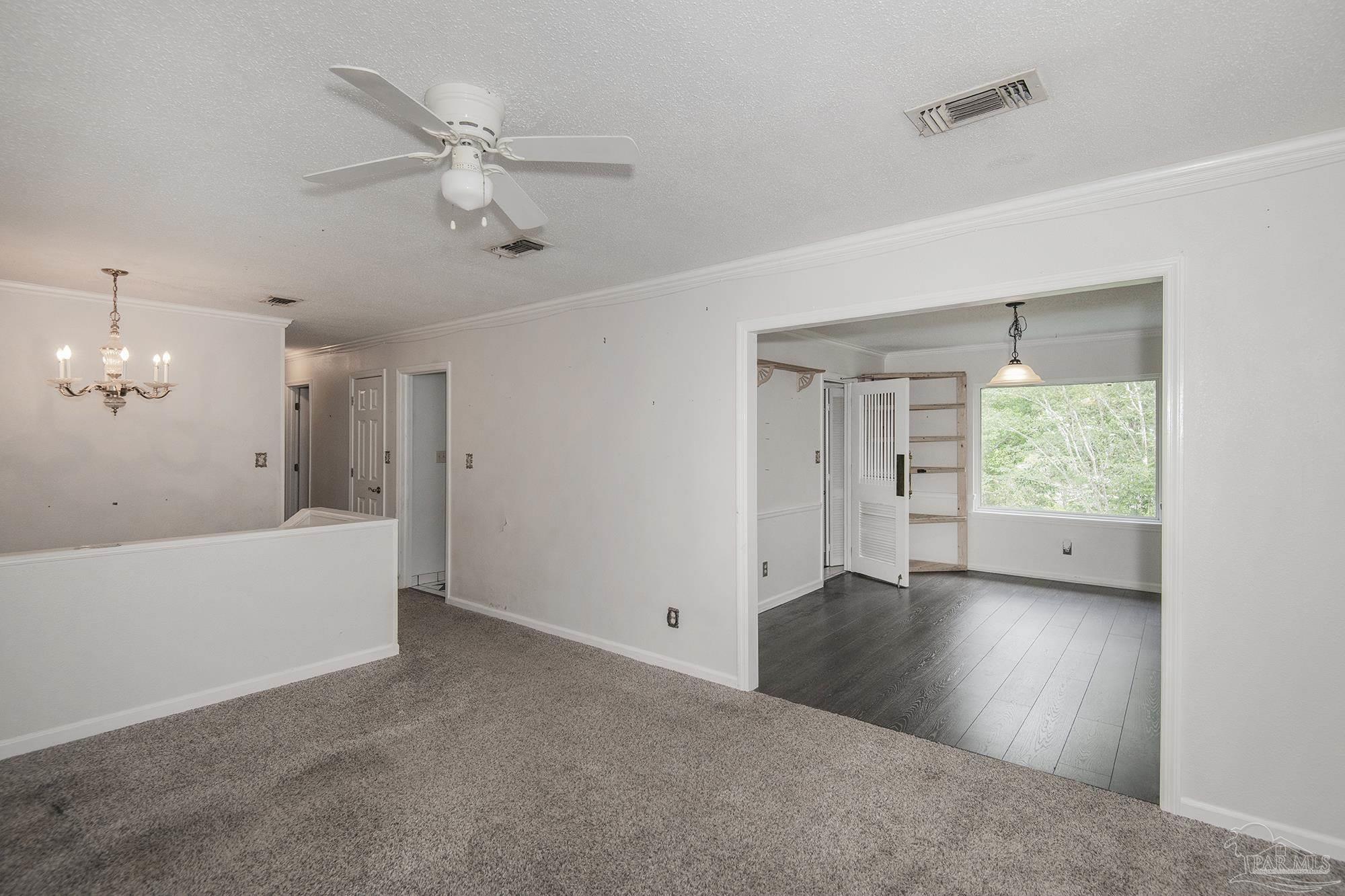 Empty carpeted room with a ceiling fan, opening to a wood-floored room with built-in shelves and a window.