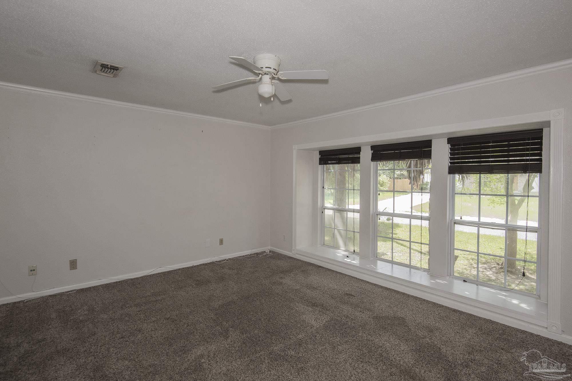 Empty carpeted room with a ceiling fan and three large windows overlooking the lawn.
