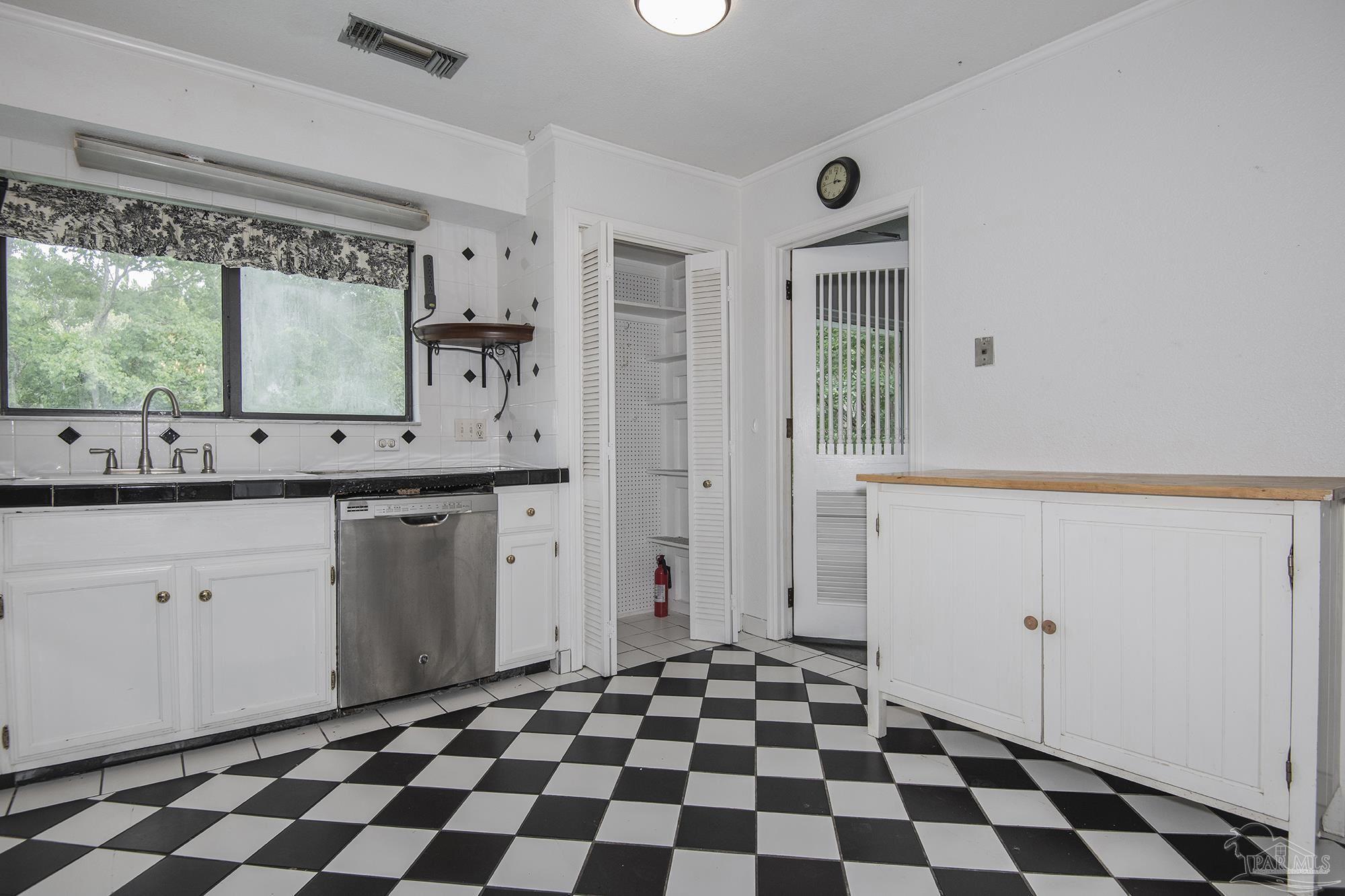 Kitchen with white cabinets, a stainless steel dishwasher, checkerboard floor, and window facing trees.