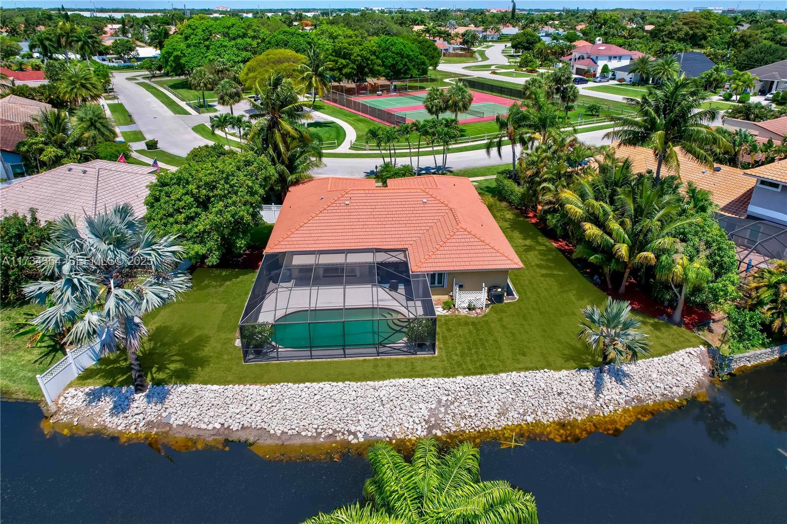 Aerial view of a canal-side home with a screened pool enclosure and nearby tennis courts.