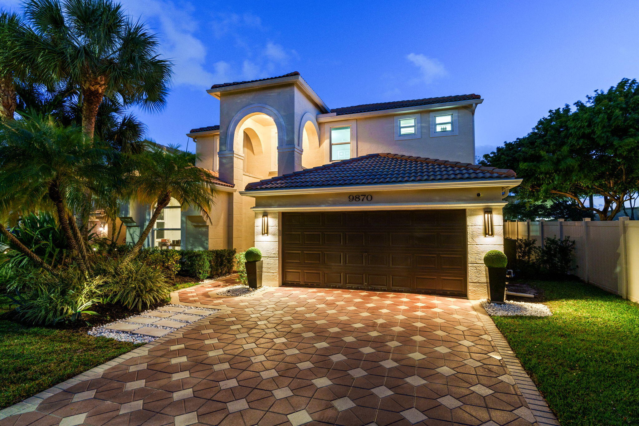 Two-story stucco home with an arched entry, double garage, and tropical landscaping at dusk.