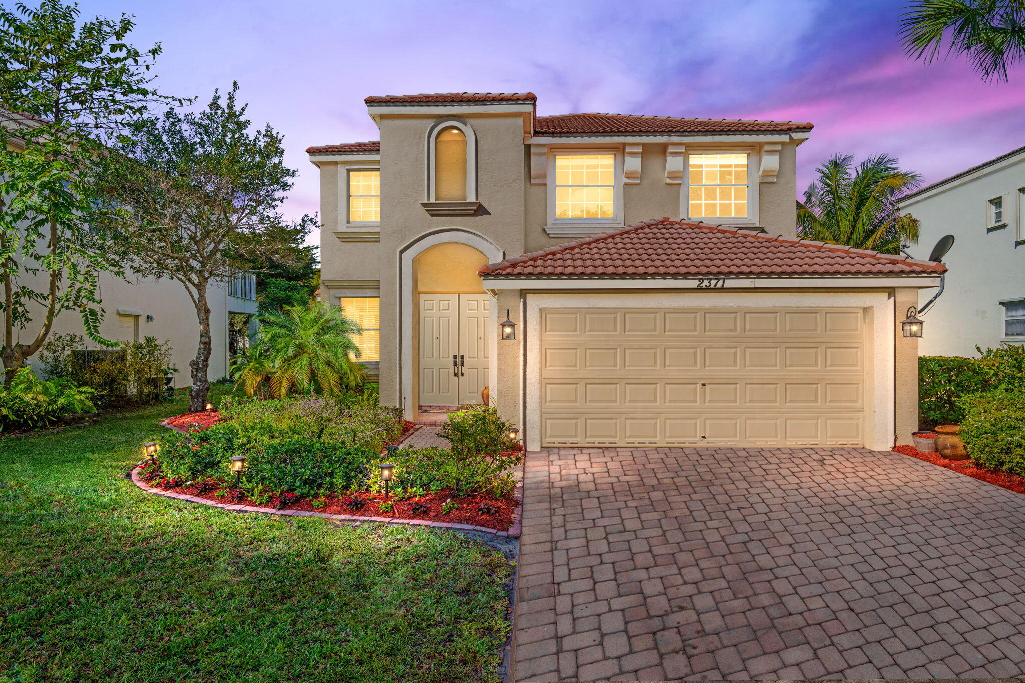 Two-story stucco home with a red tile roof, arched entry, and wide paved driveway at dusk.