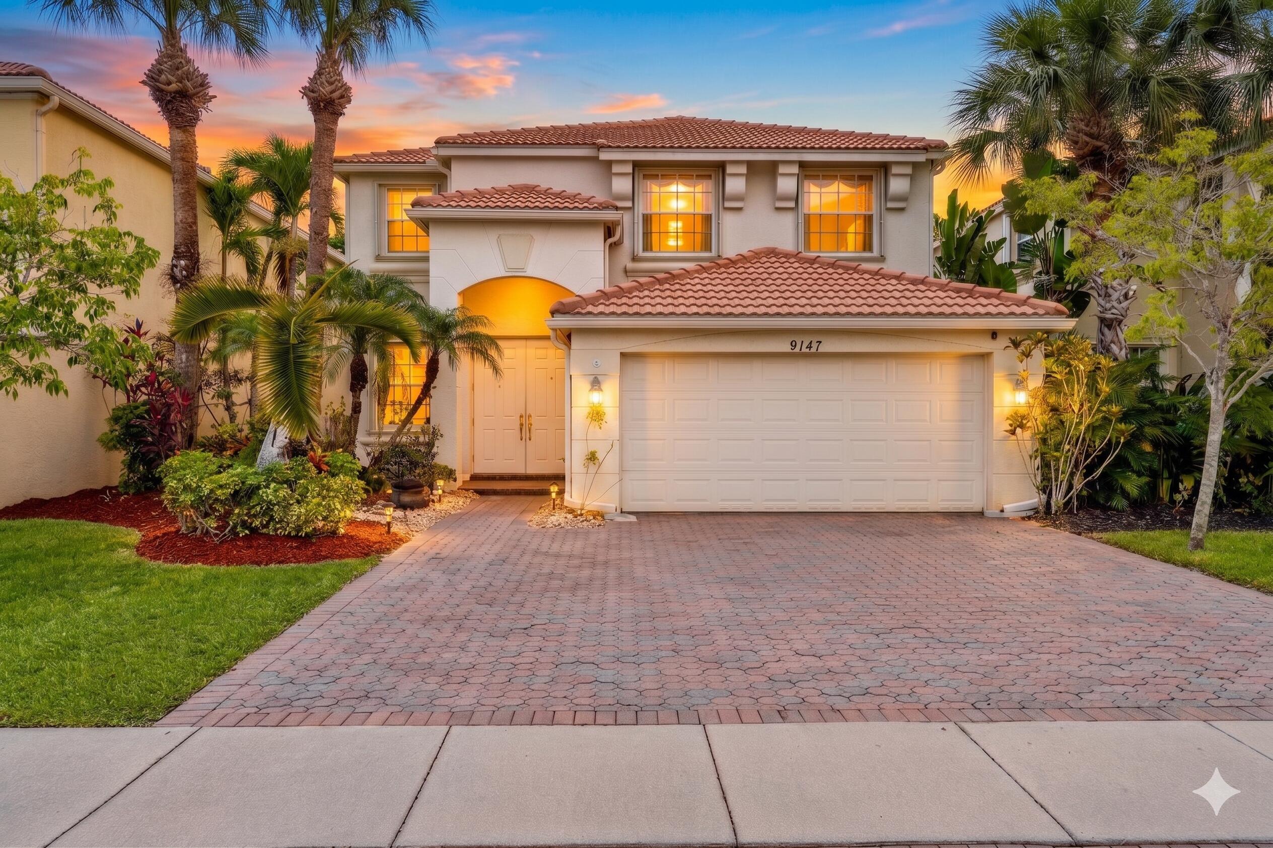 Two-story stucco home with a red tile roof, glowing windows, and tropical landscaping at dusk.