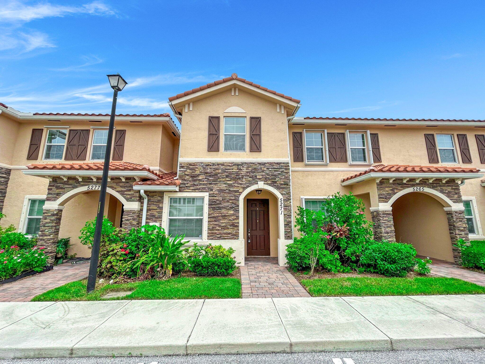 Two-story beige townhouse with stone accents, arched entry, tiled roof, and landscaped front walkway.