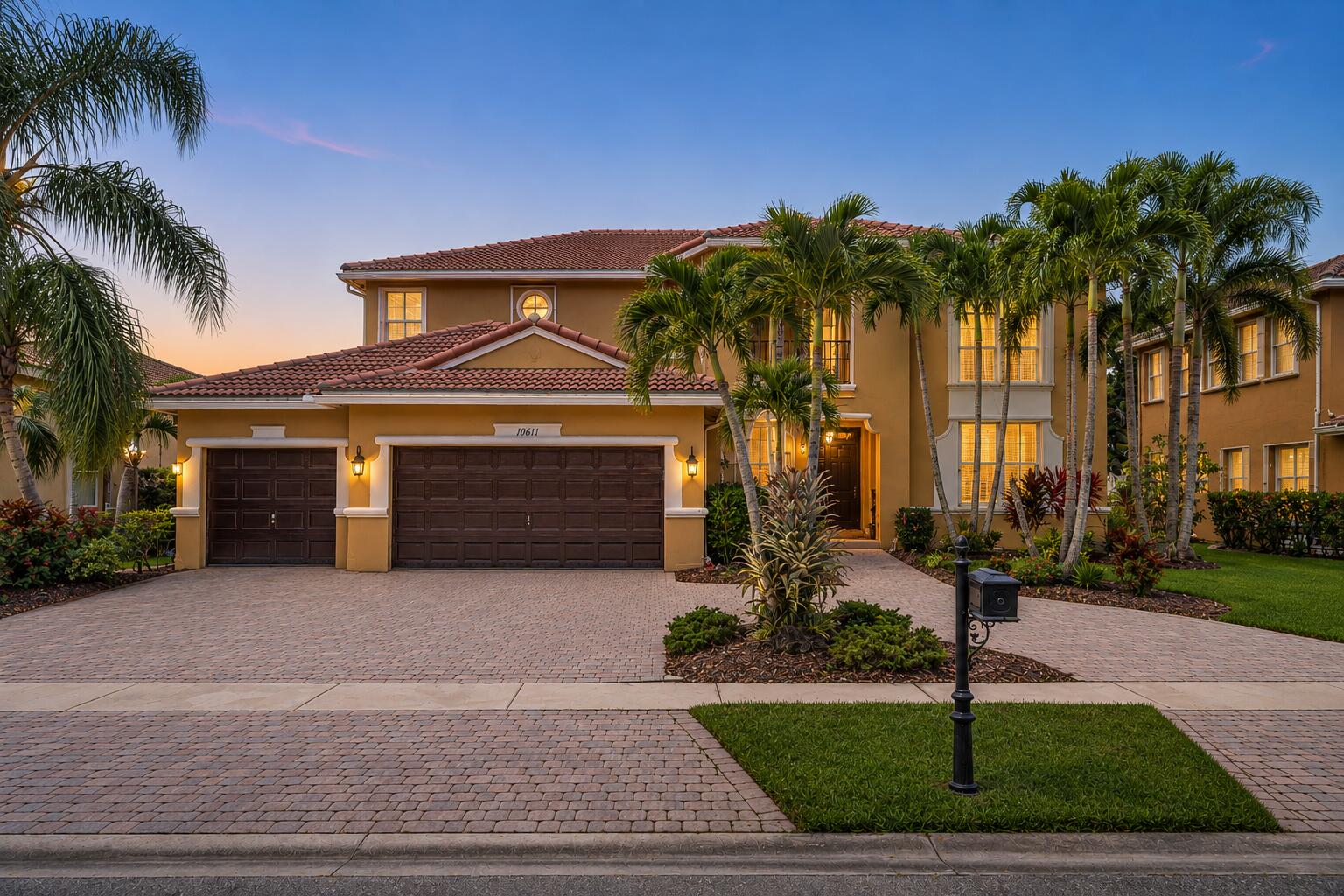 Two-story tan stucco home with a red tile roof, palm trees, and a wide paver driveway at dusk.