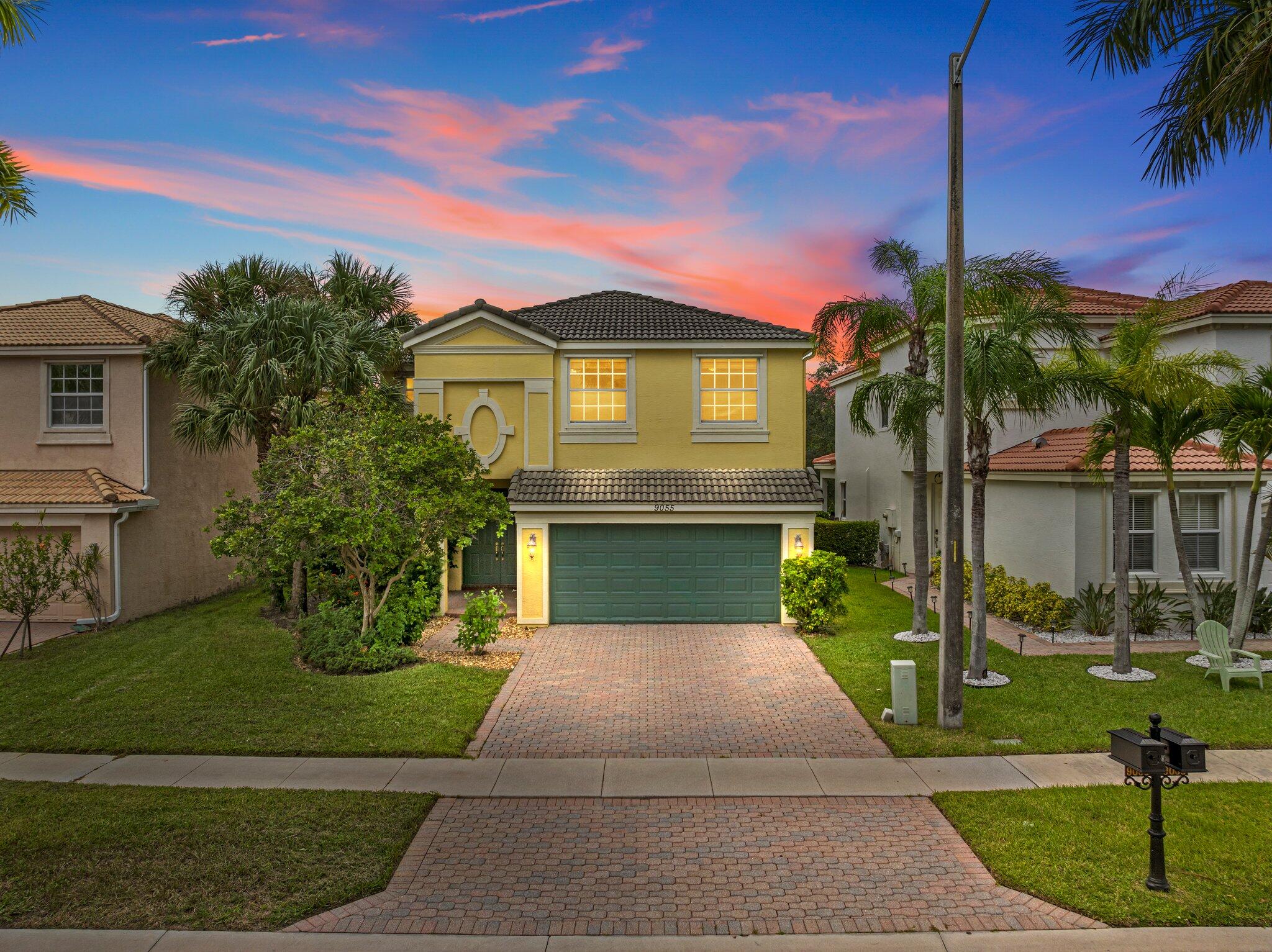 Yellow two-story home with a green double garage door and brick-paver driveway at dusk.