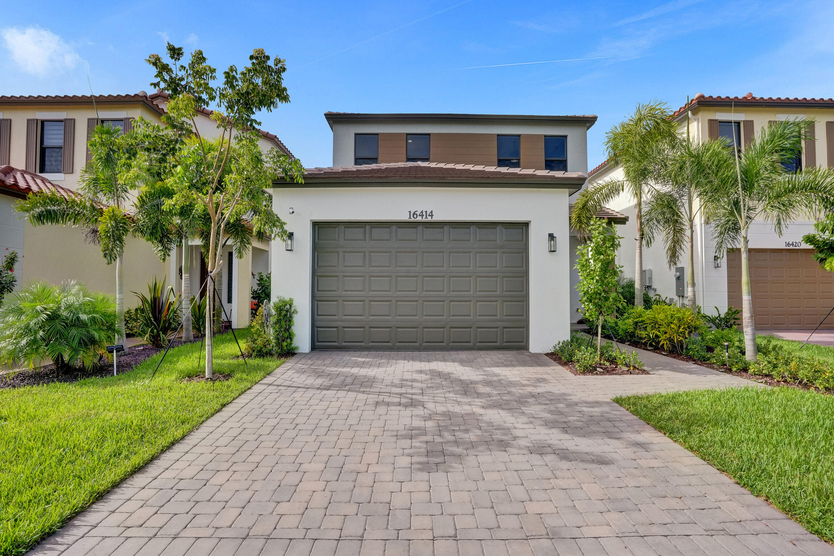 Modern white two-story home with a dark garage, broad paver driveway, and tropical landscaping.