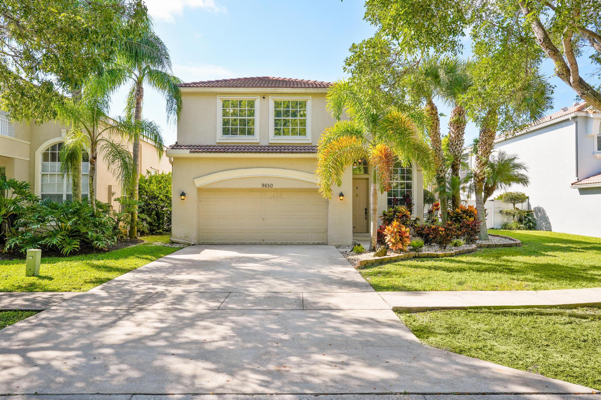 Two-story cream stucco home with a red tile roof, double garage, and tropical palms around the driveway.