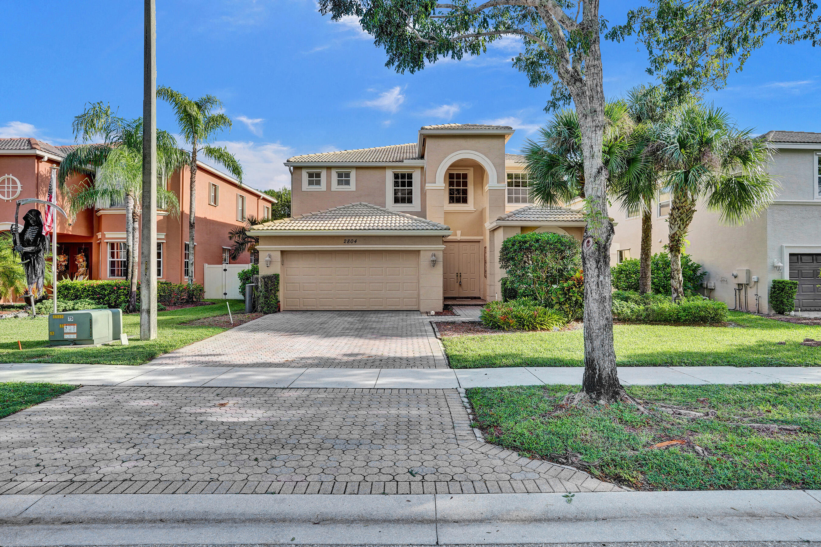 Two-story beige stucco home with an arched window, double garage, and palm-lined paver driveway.
