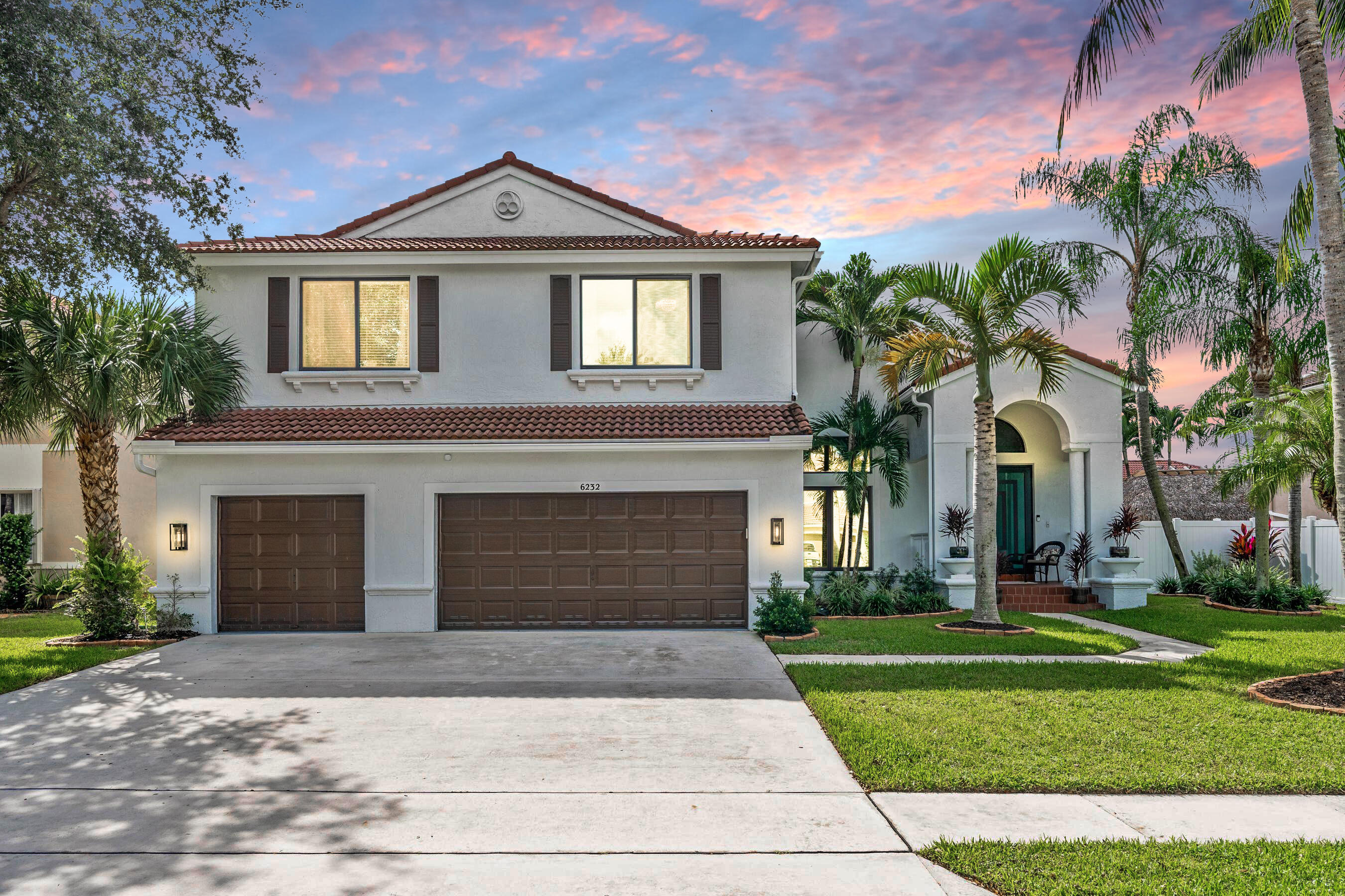 Two-story stucco home with a tiled roof, double garage, arched entry, and palm-lined landscaping at dusk.