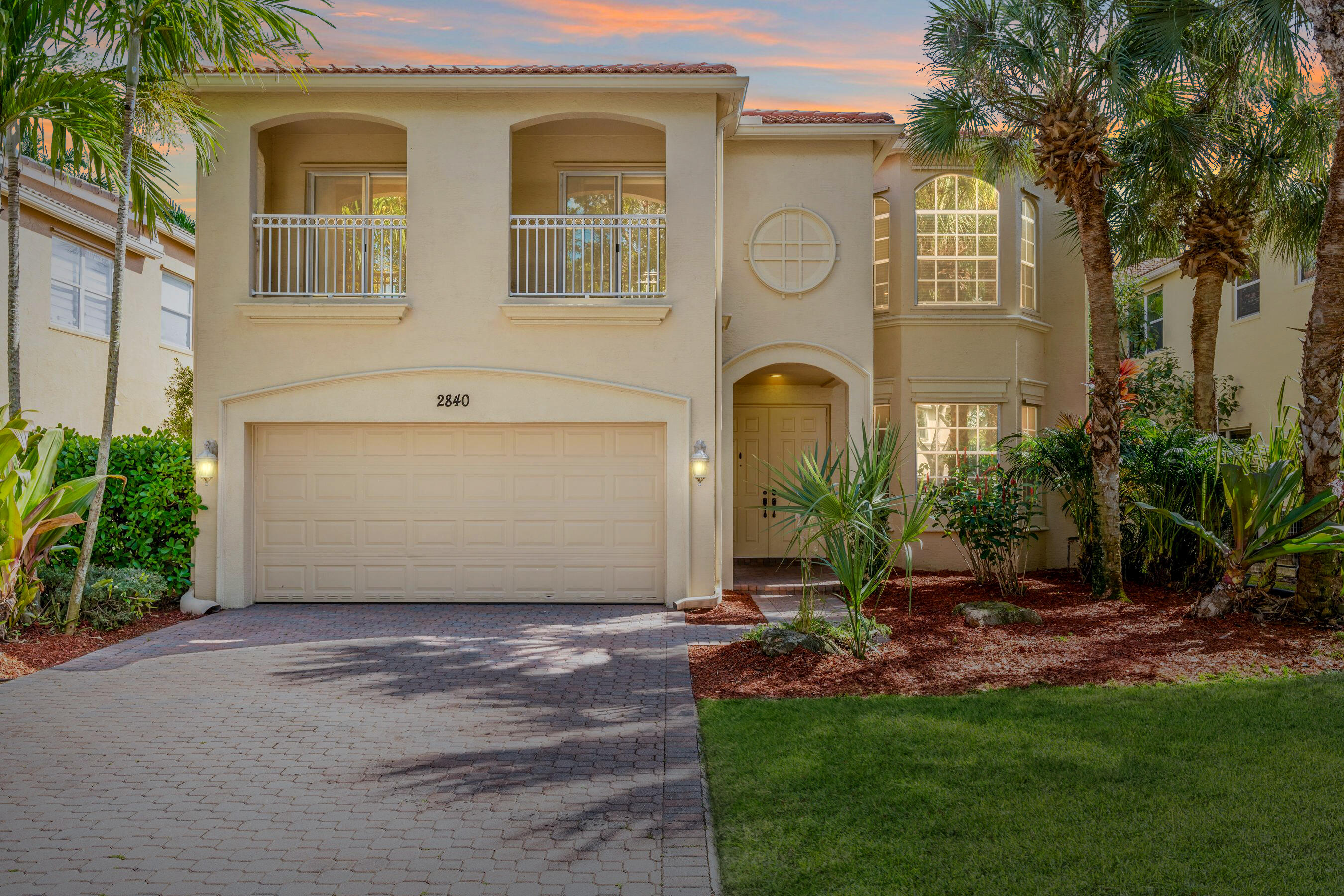 Two-story stucco home with palm trees, a paver driveway, and a two-car garage.