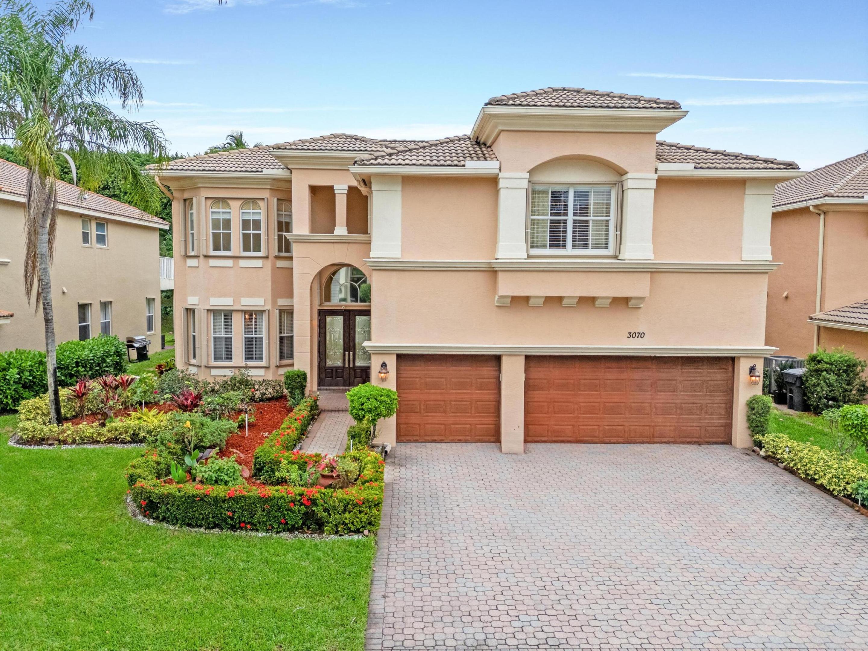 Two-story peach stucco home with a tiled roof, double garage, paver driveway, and landscaped garden.