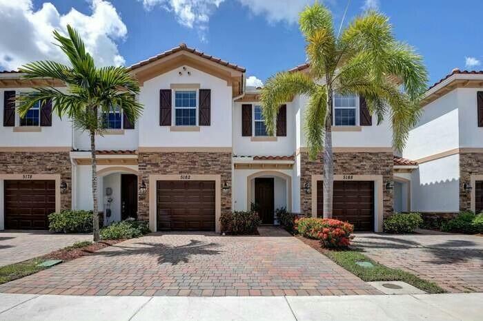 Two-story townhomes with stone accents, brown garage doors, palm trees, and brick driveways.