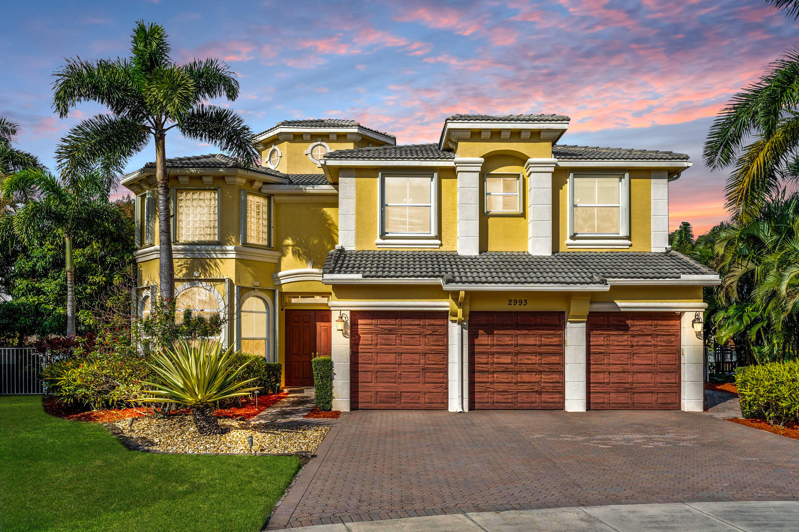 Yellow two-story house with three wooden garage doors, framed by palm trees at dusk.