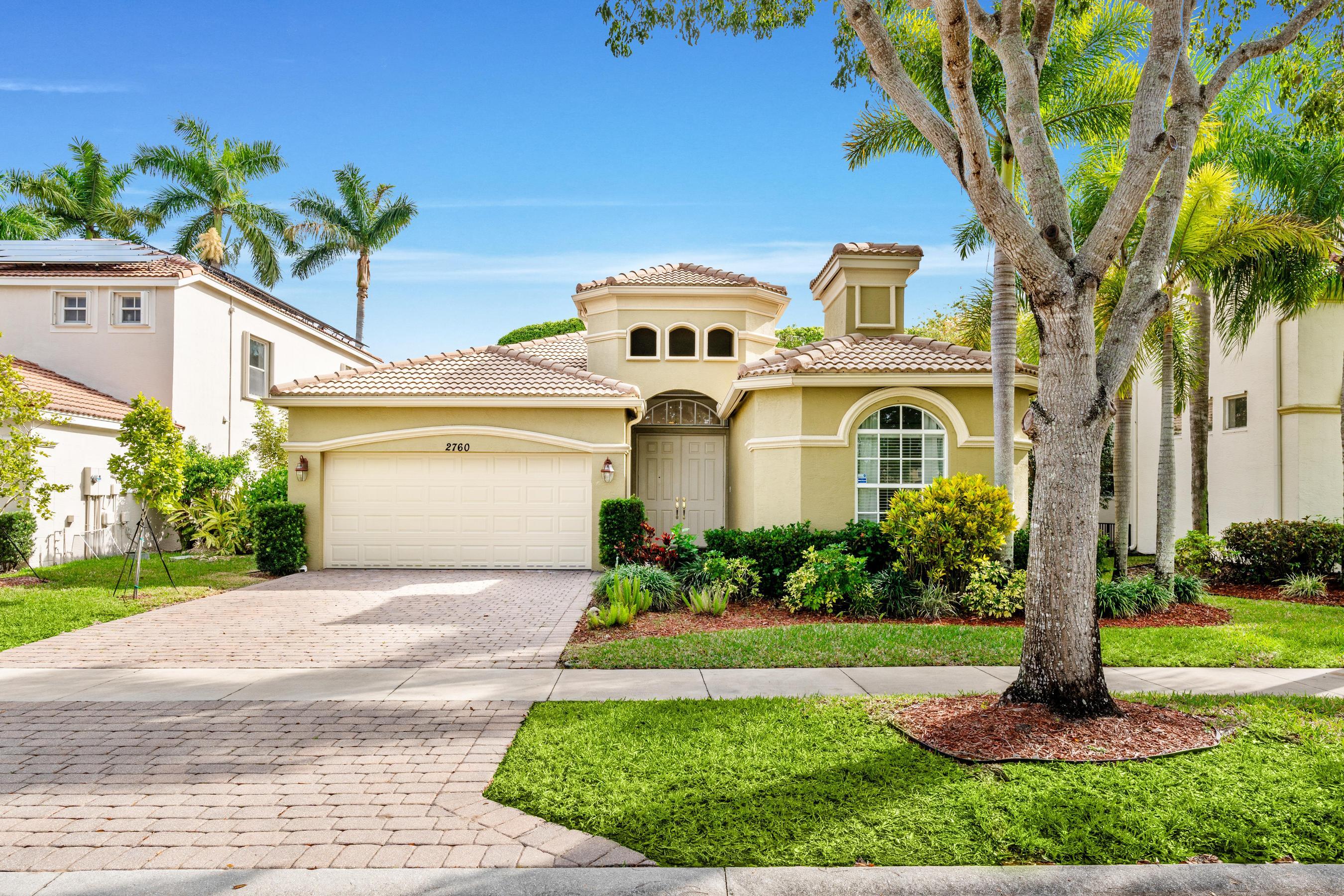 Cream stucco home with a tiled roof, arched window, double garage, and tropical landscaping.