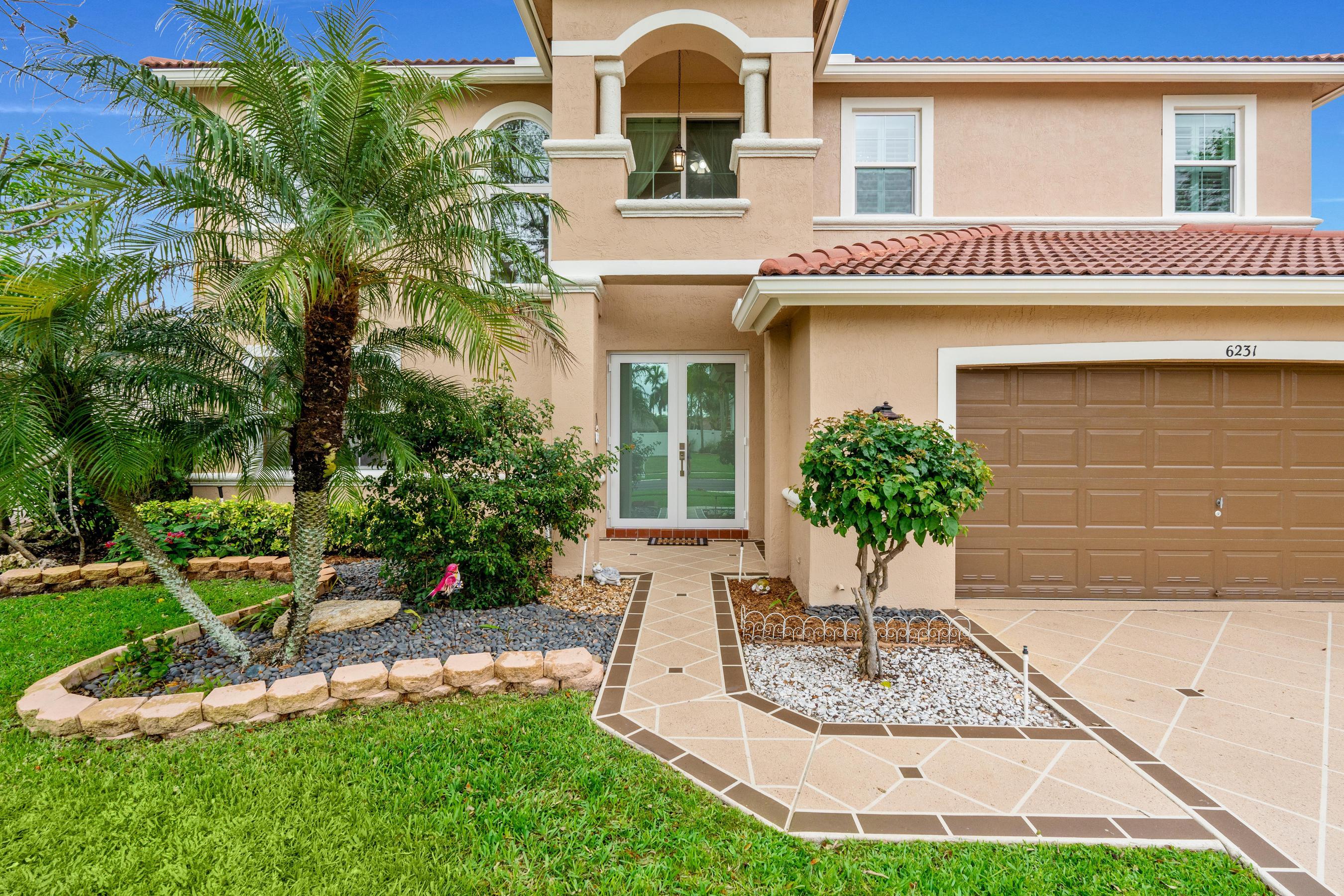 Two-story stucco home with terracotta roof, tiled entry walkway, brown garage door, and tropical landscaping.