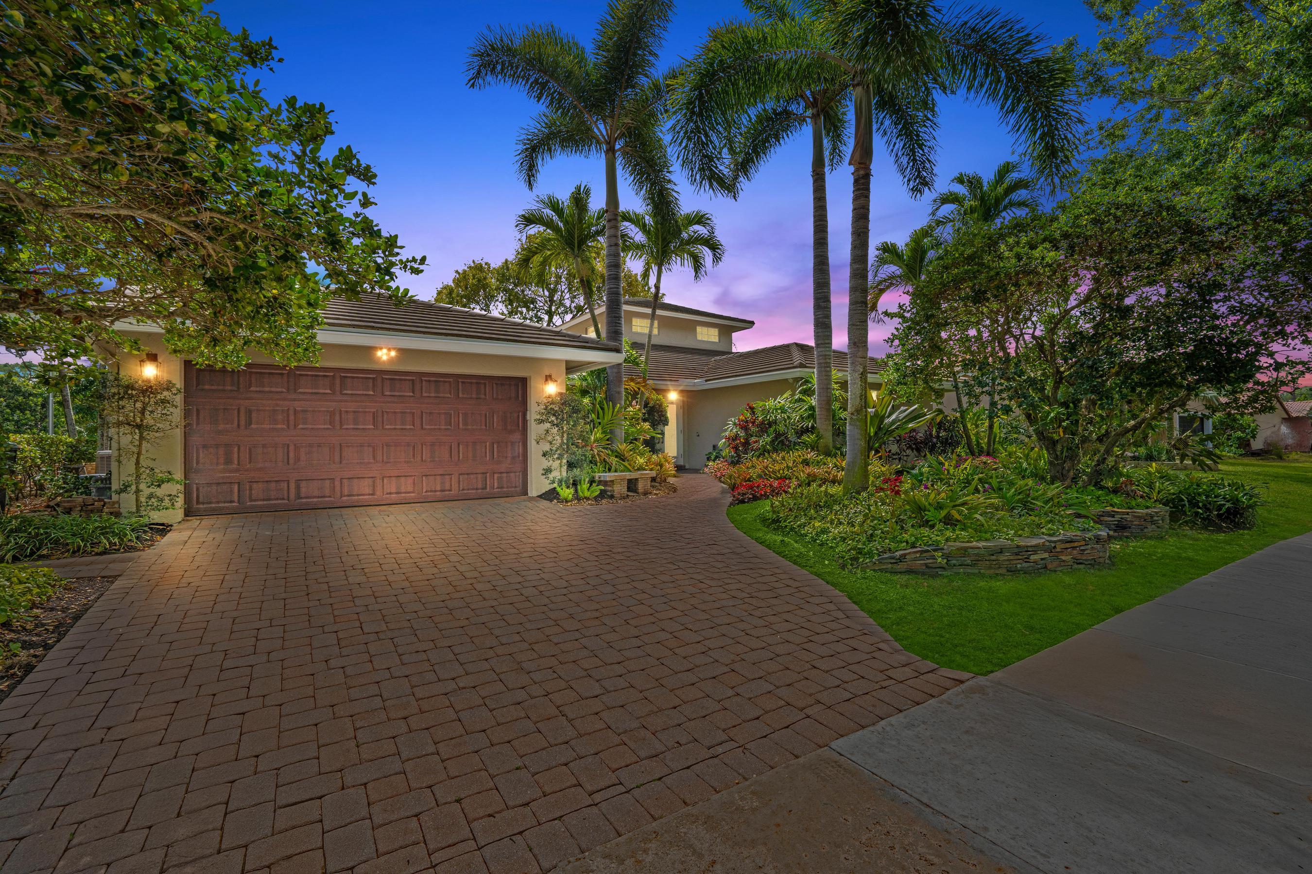 Two-story home with a broad paver driveway, wooden garage door, and tropical landscaping at dusk.