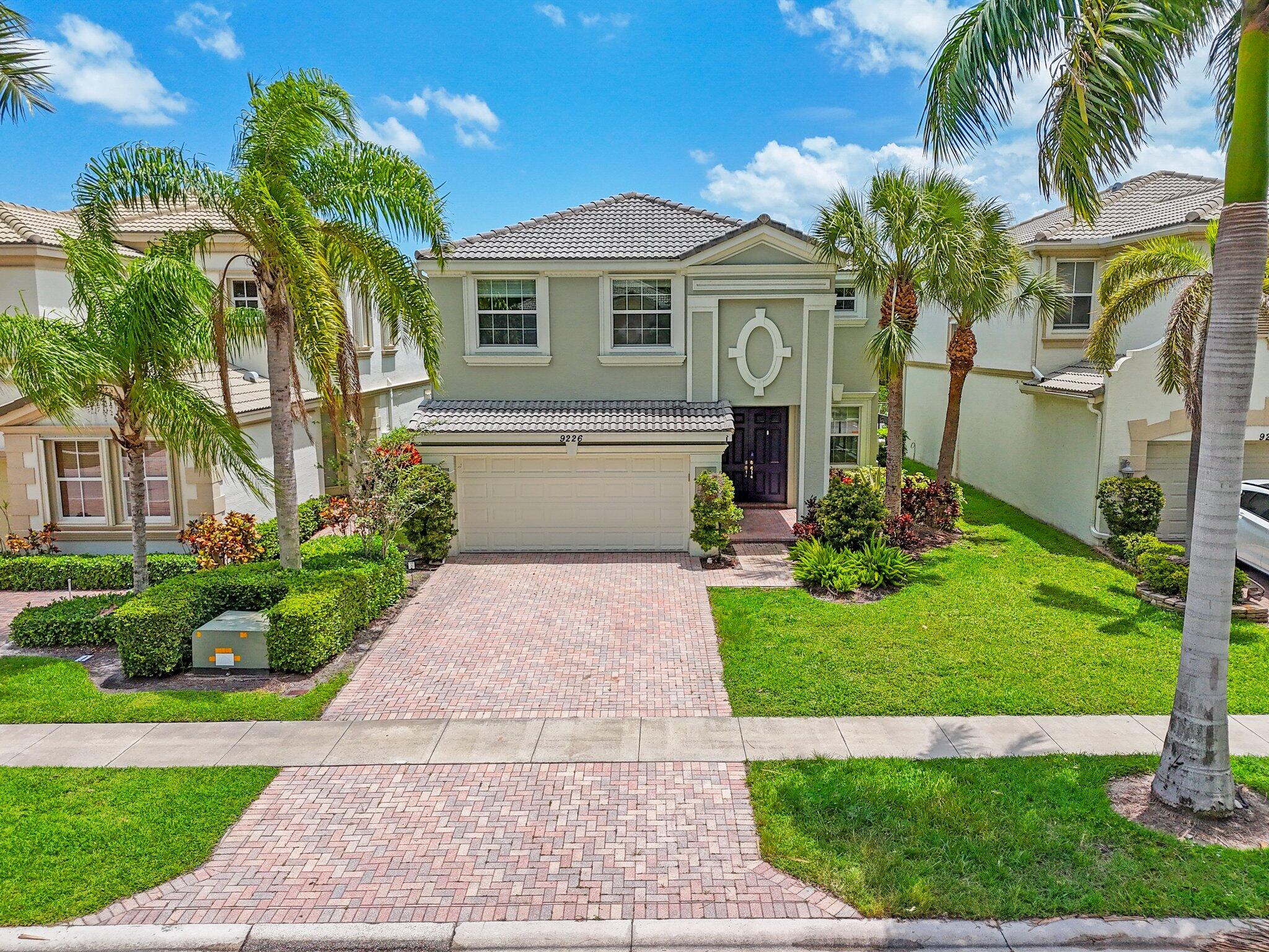 Two-story stucco home with a tiled roof, brick driveway, two-car garage, and palm-lined lawn.