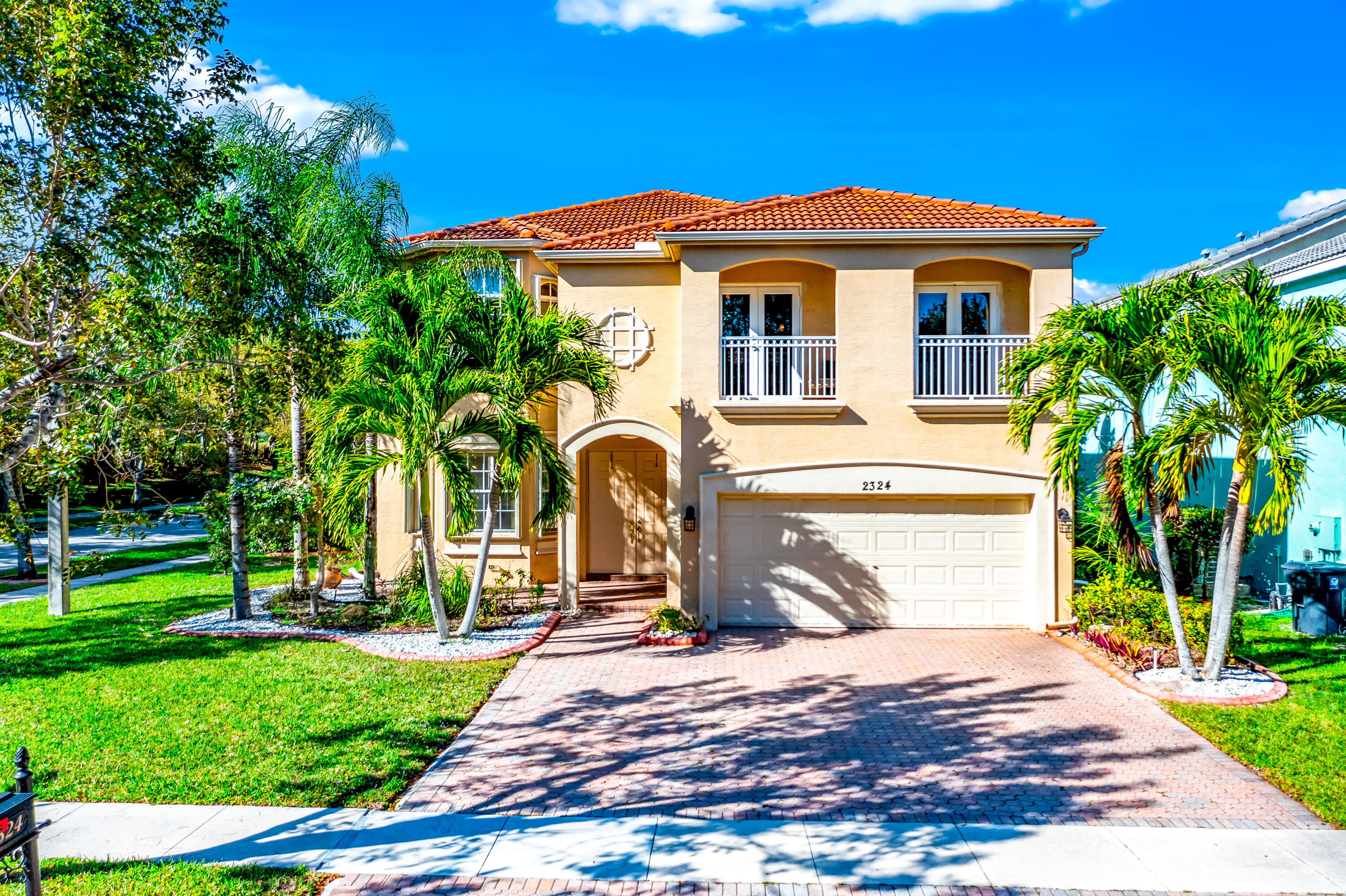 Two-story stucco house with a terracotta roof, upper balconies, double garage, and palm-lined driveway.
