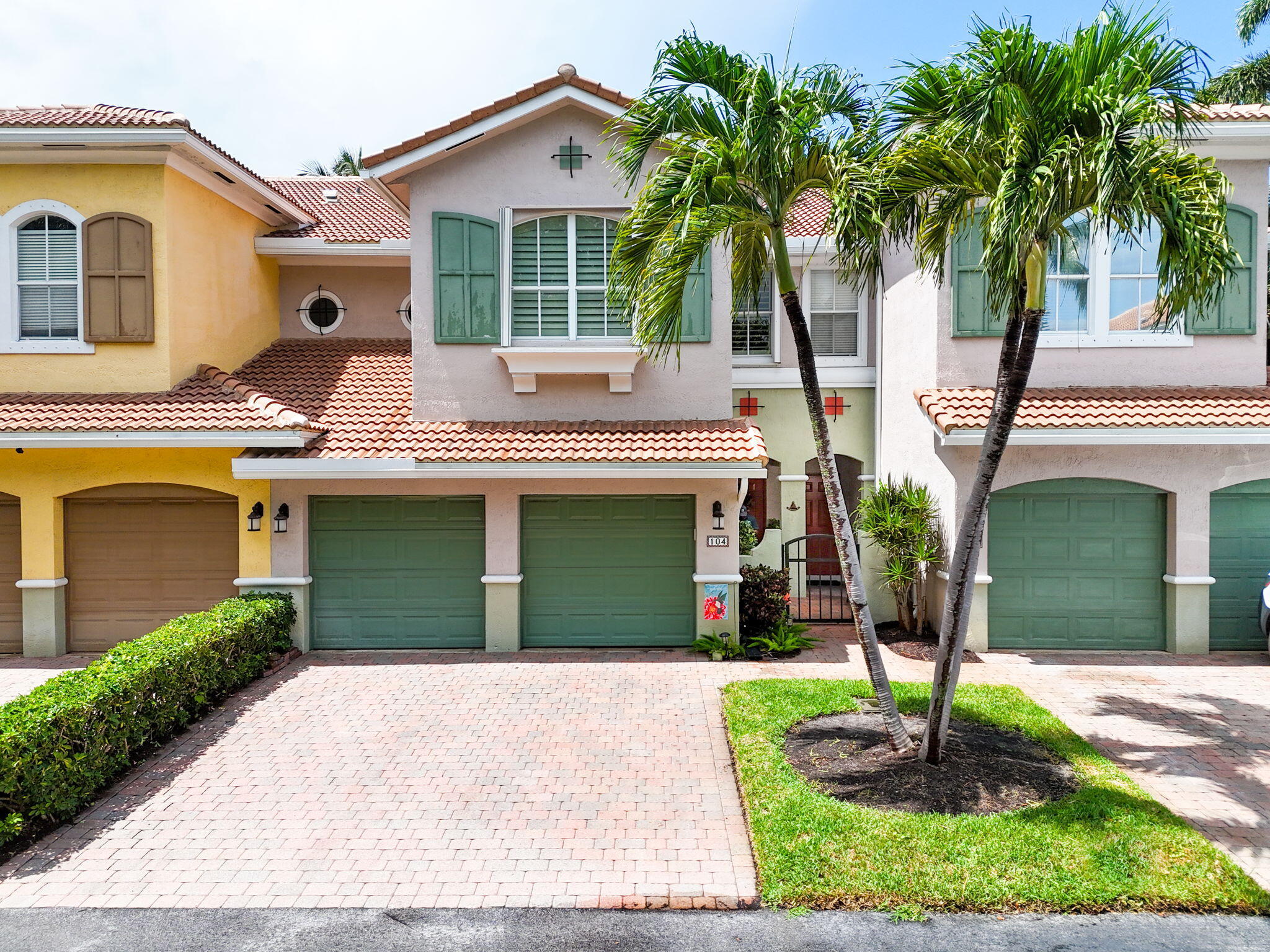 Pastel stucco homes with terracotta roofs, green garage doors, and palm trees beside brick-paver driveways.
