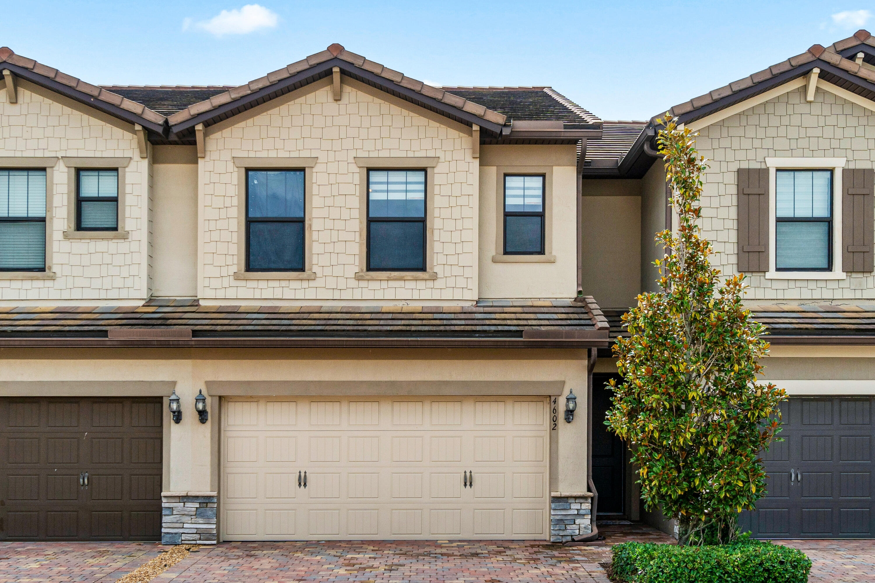 Cream-colored multi-unit home with several garage doors and a leafy tree beside the entrance.