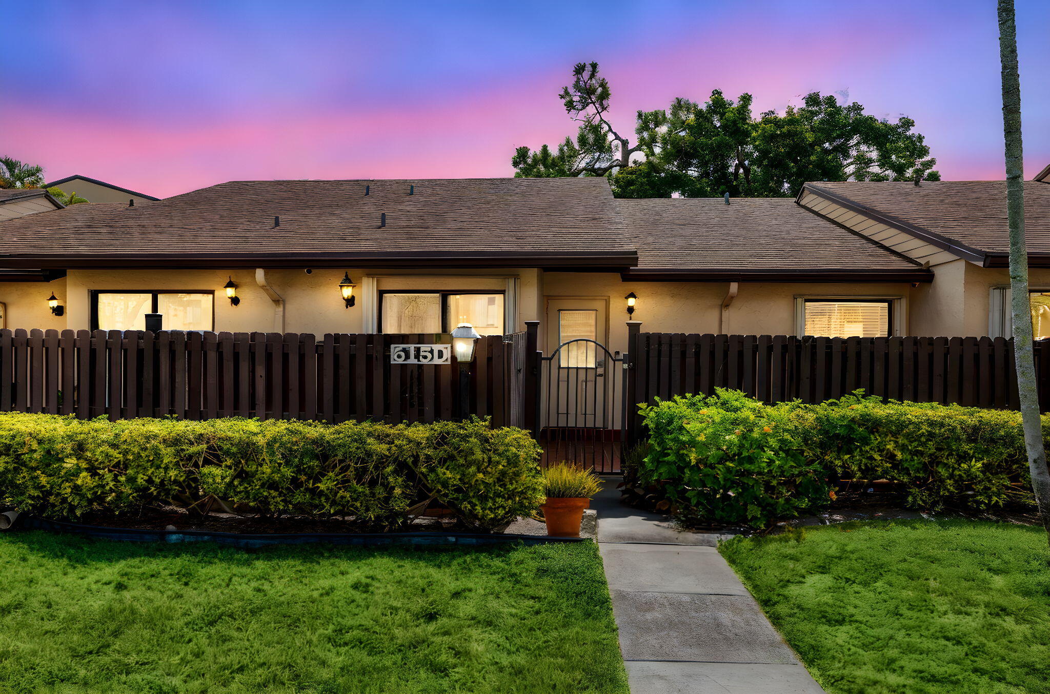 Single-story home behind a dark wooden fence, with a walkway, trimmed hedges, and glowing exterior lights.