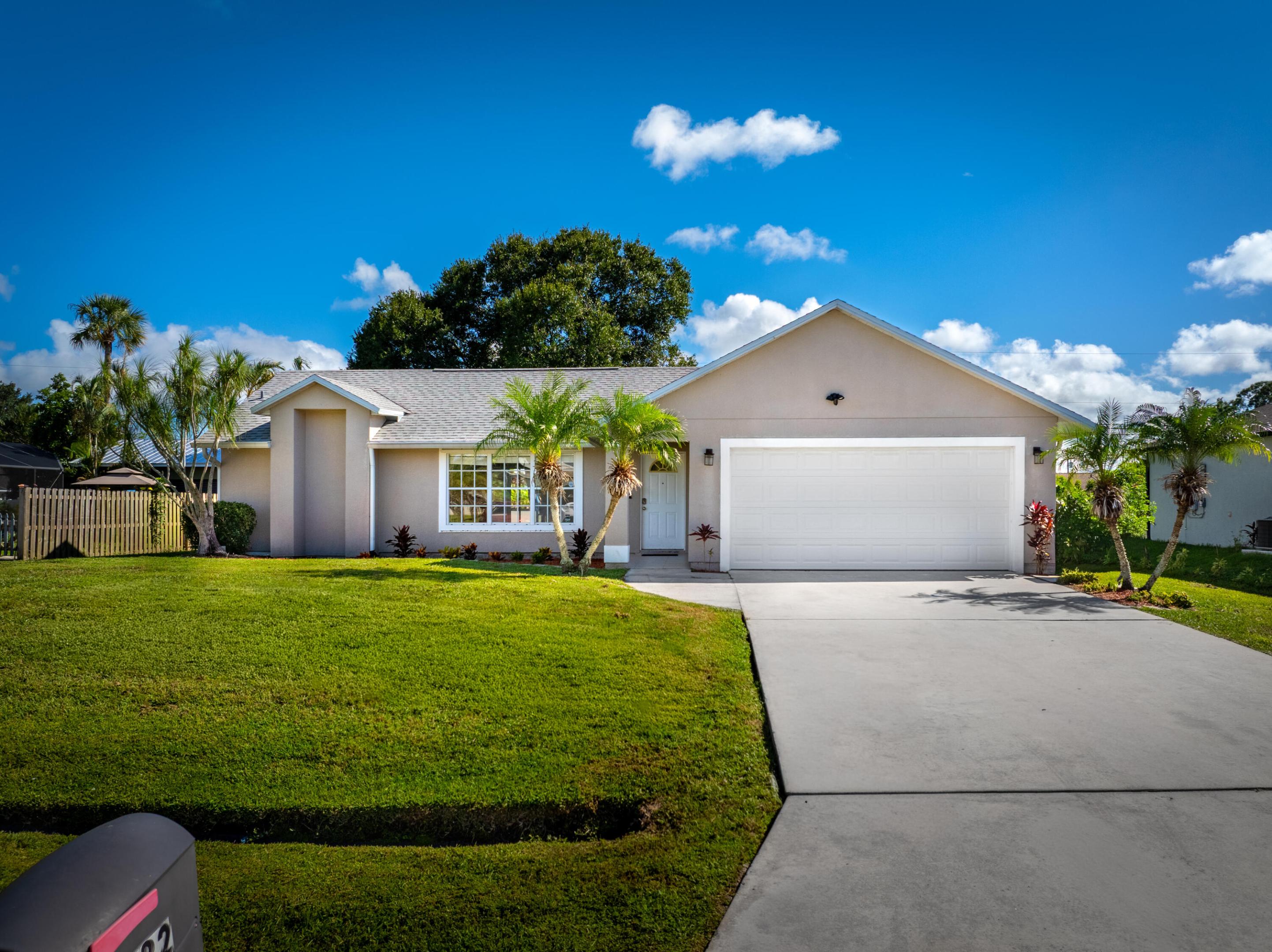 Single-story beige home with a white garage, palm trees, green lawn, and wide driveway.