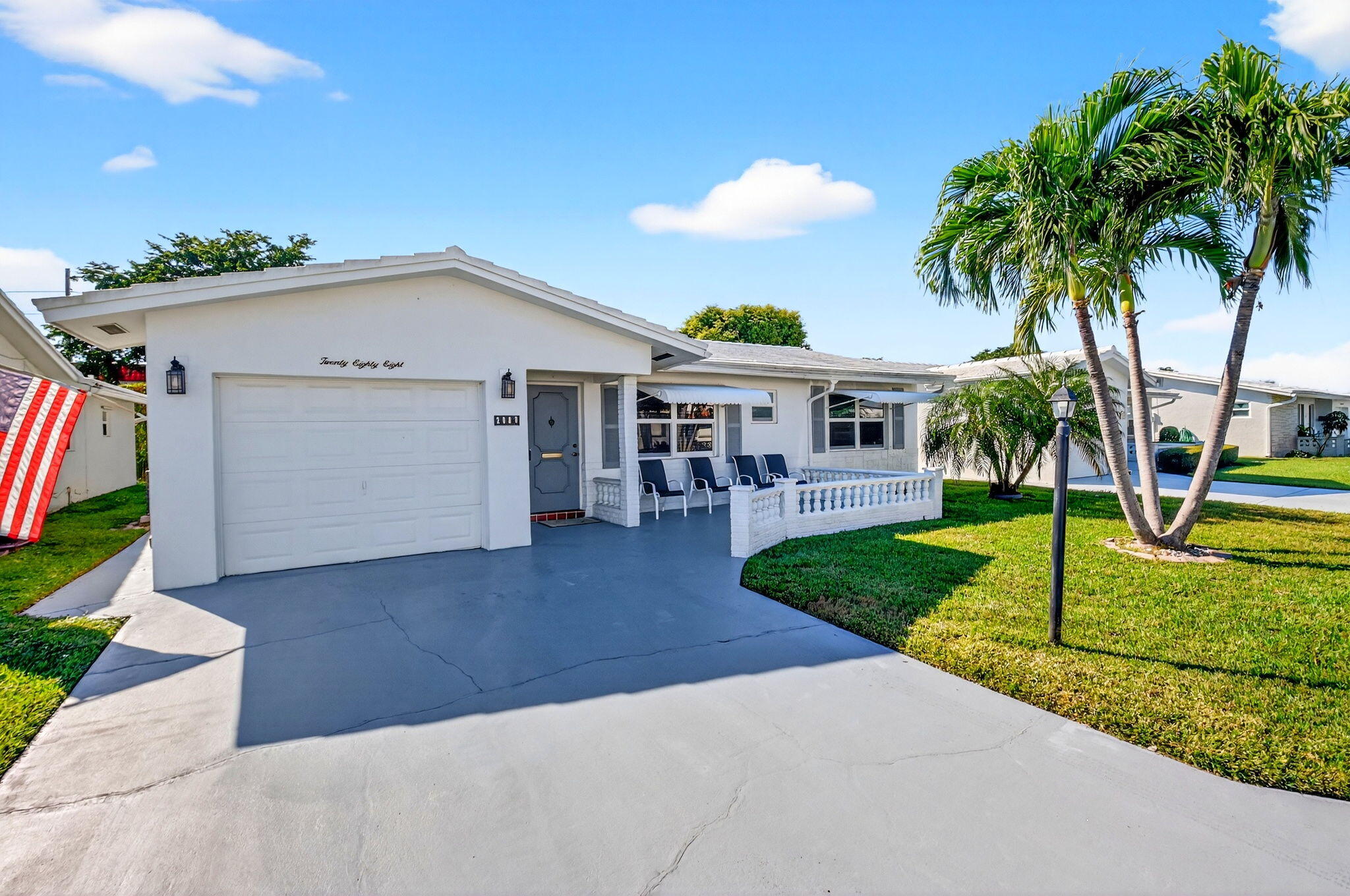 White single-story home with a covered porch, broad driveway, green lawn, and tall palm trees.