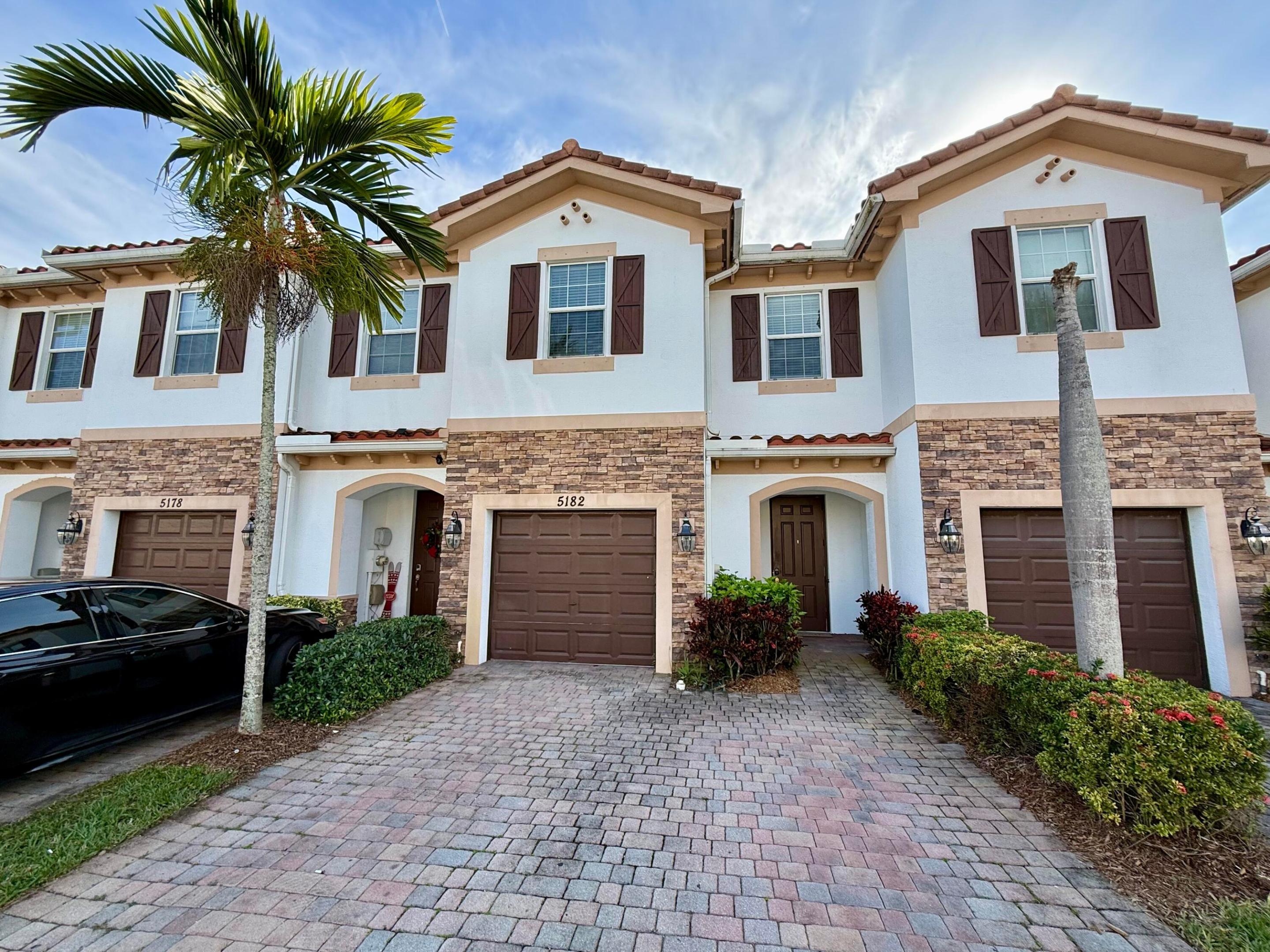 Two-story townhouses with stone façades, brown shutters, palm trees, and a paved driveway.