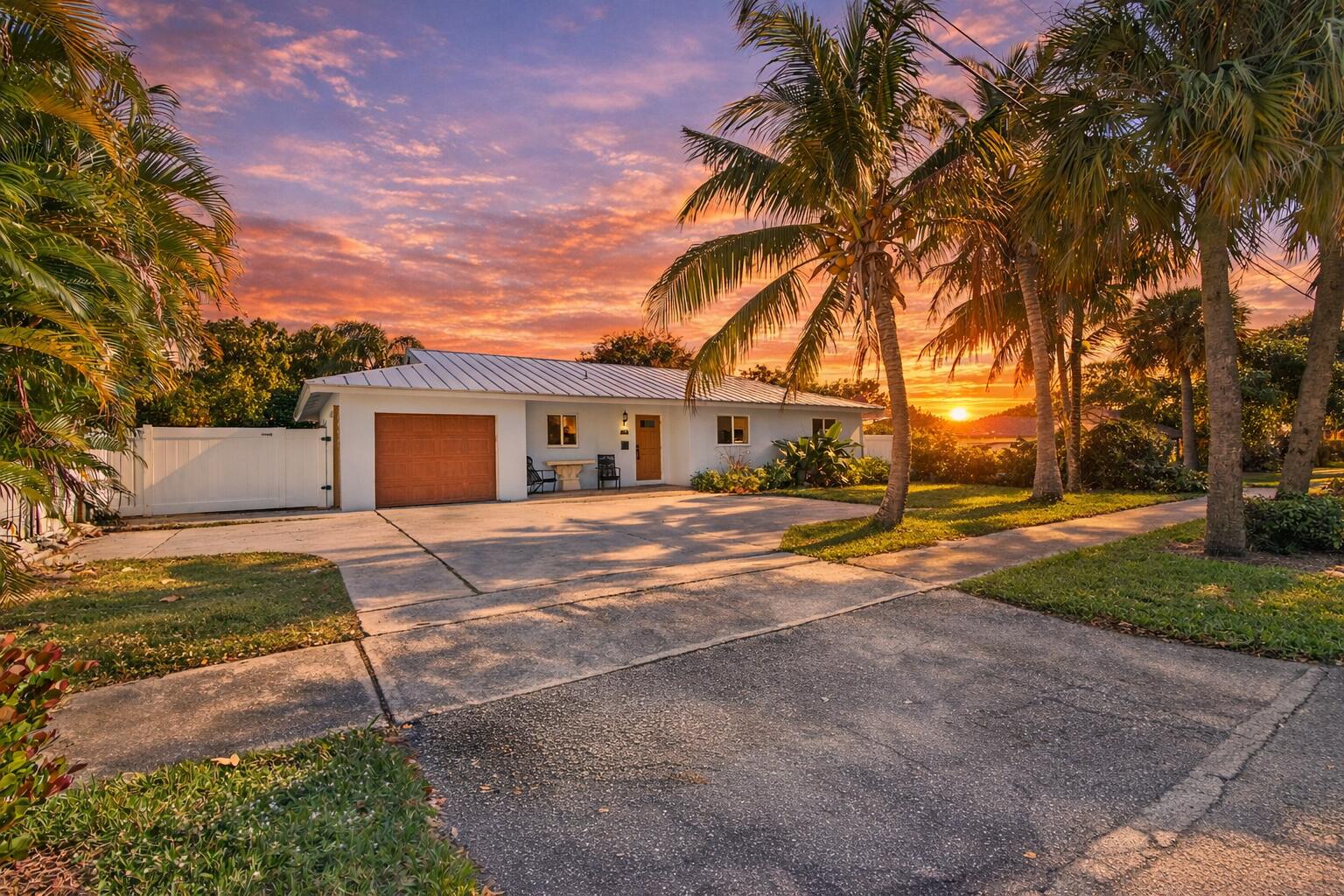 Single-story white home framed by palm trees beneath a vivid sunset, with water visible behind it.