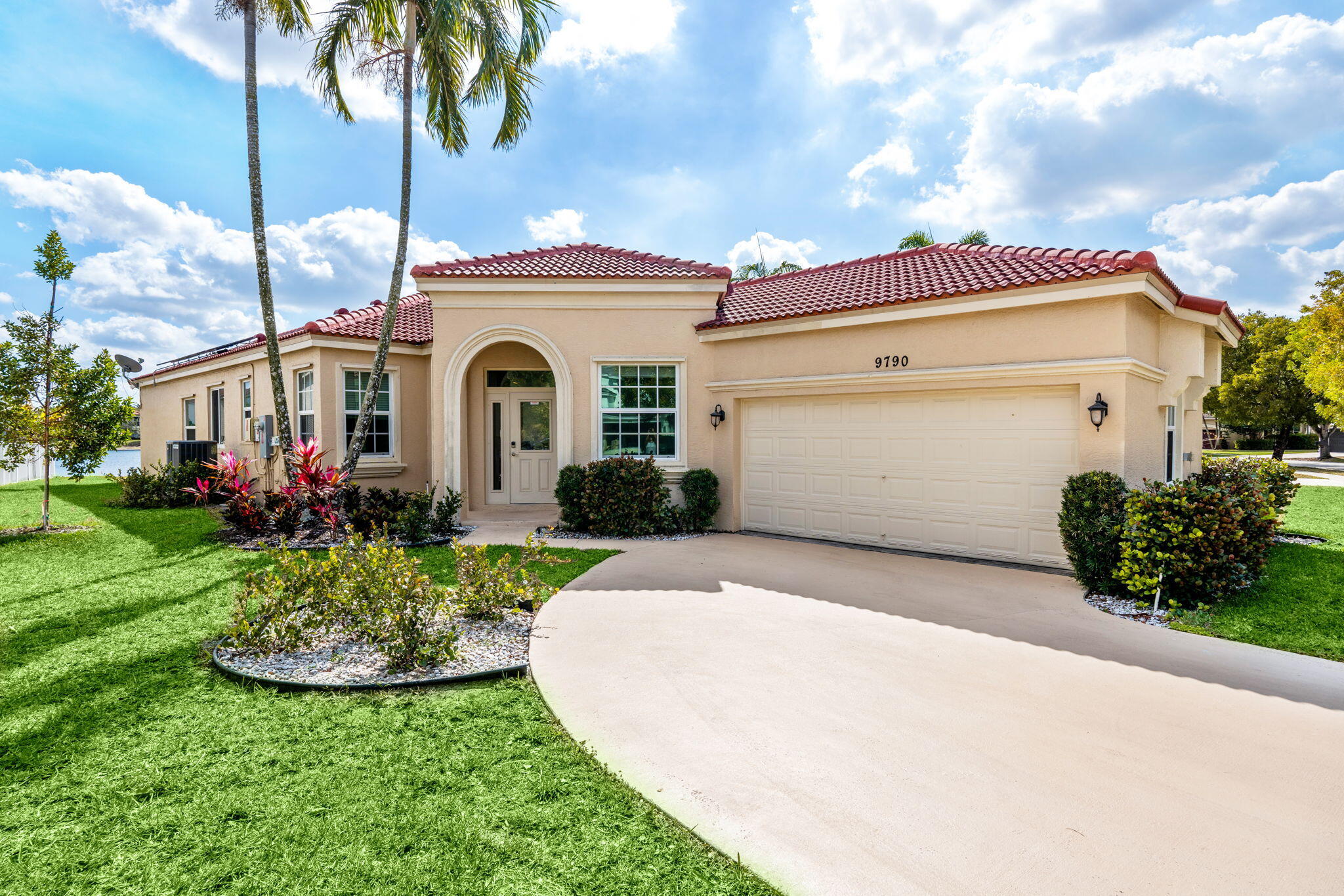 Beige stucco home with a red tile roof, arched entry, two-car garage, and palm-lined lawn.