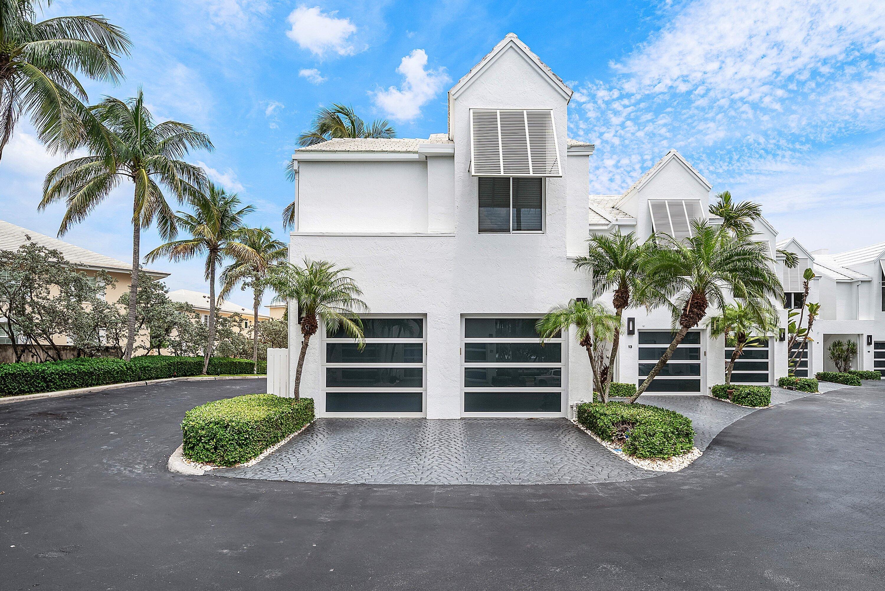 White two-story townhouse with dark garage doors, palm trees, and a paved driveway.